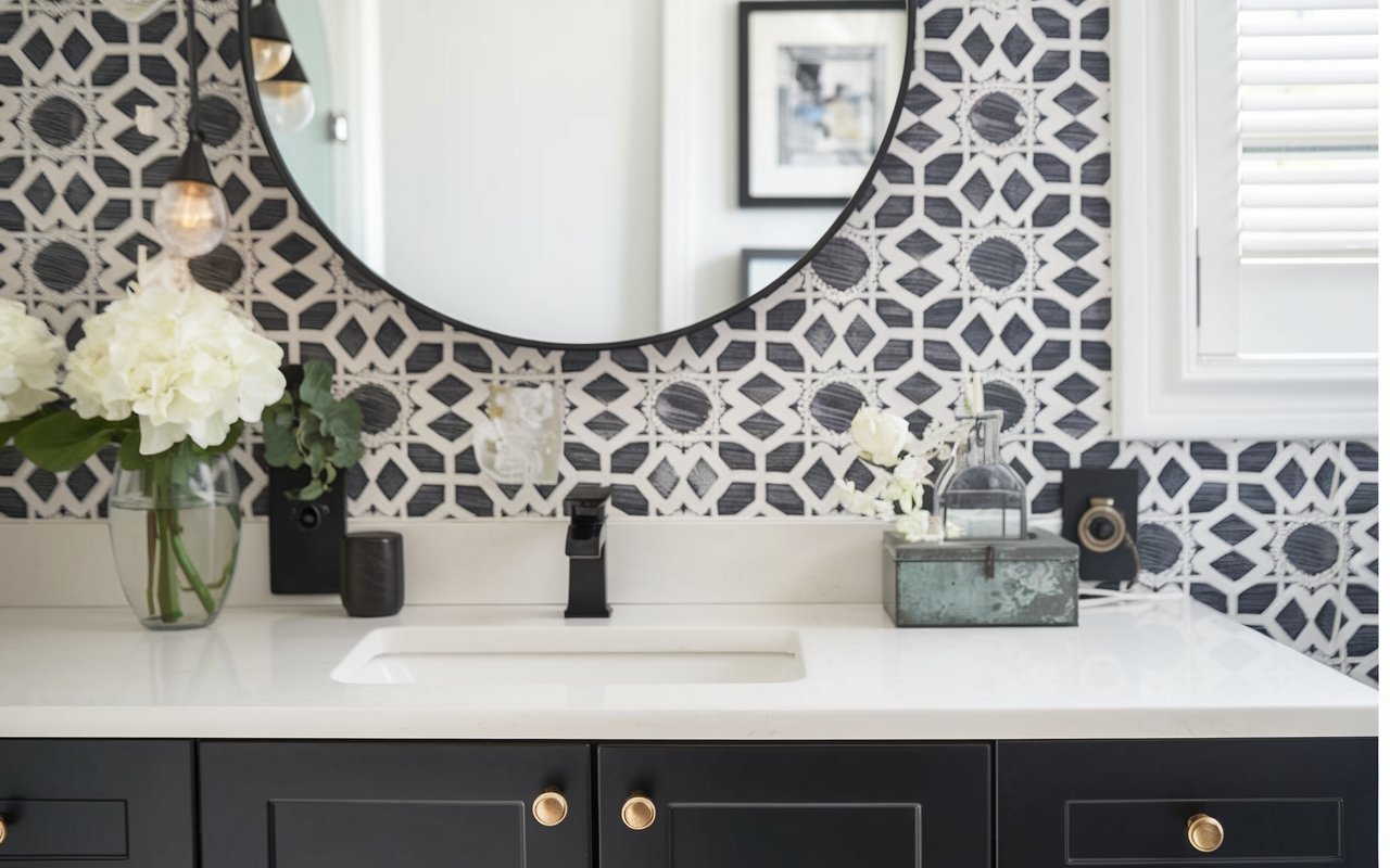 Modern bathroom vanity featuring a round mirror and geometric black-and-white tile backsplash. With a design that embraces elegance, it includes a black faucet, a vase of white flowers, and framed photos. Bright natural lighting enhances this stylish half bathroom decor.