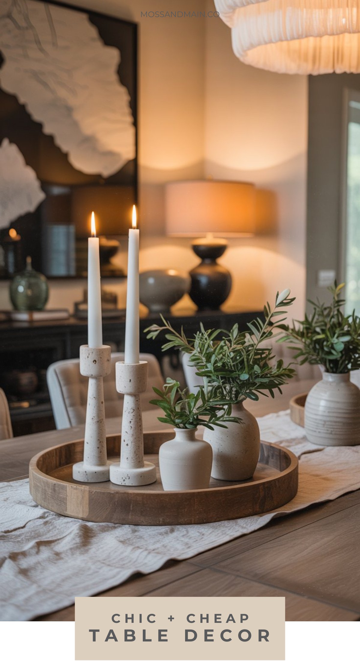 A wooden tray on a dining room table holds two lit white candles and small ceramic vases with greenery. The background features a lamp, wall art, and more plants, offering organic chic table decor ideas for a cozy, stylish space.