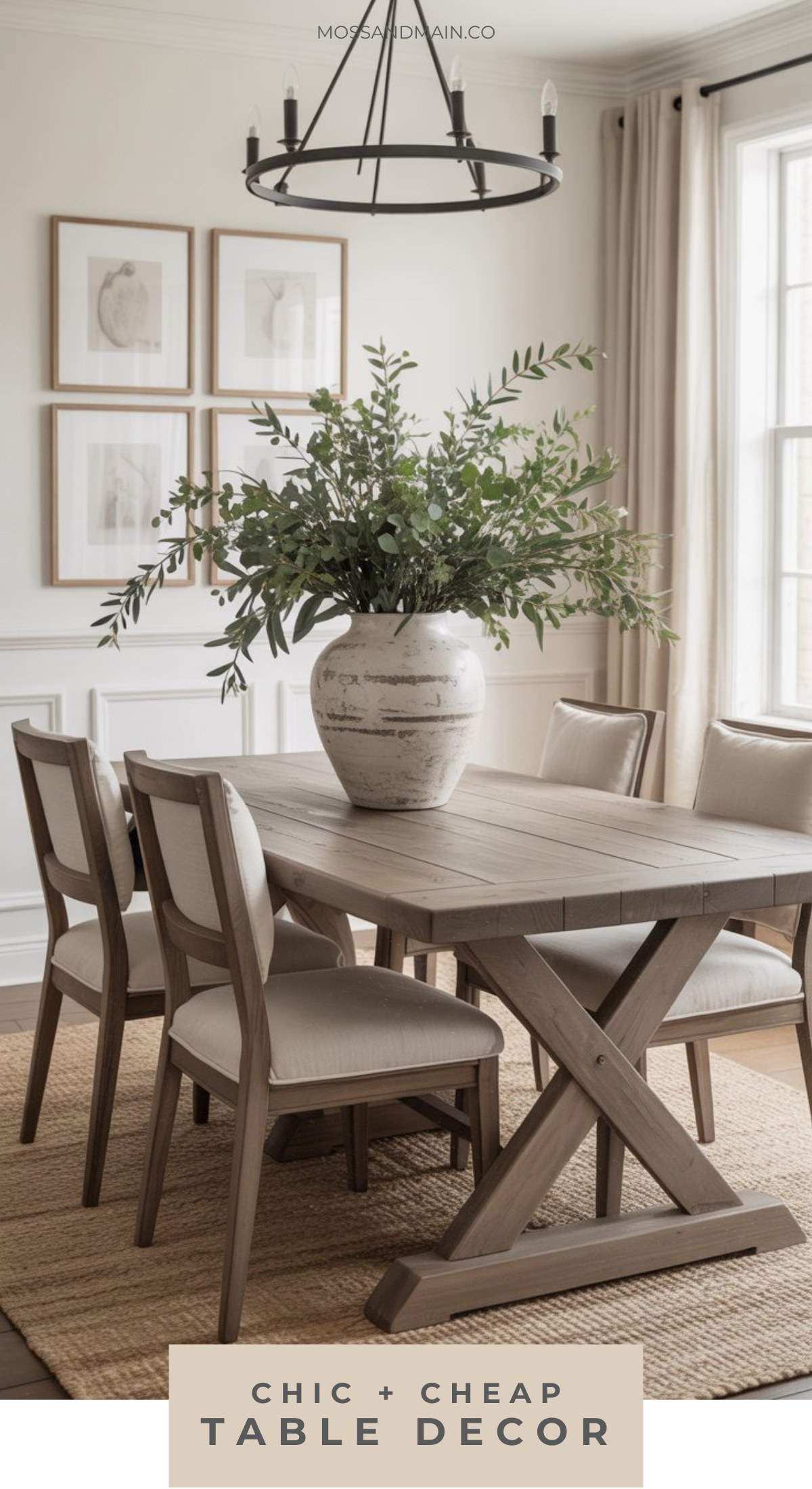 A rustic dining room with a wooden table, six beige cushioned chairs, organic chic dining room table decor, a large white vase with green foliage, neutral wall art, black chandelier, and natural light from cream-curtained windows.