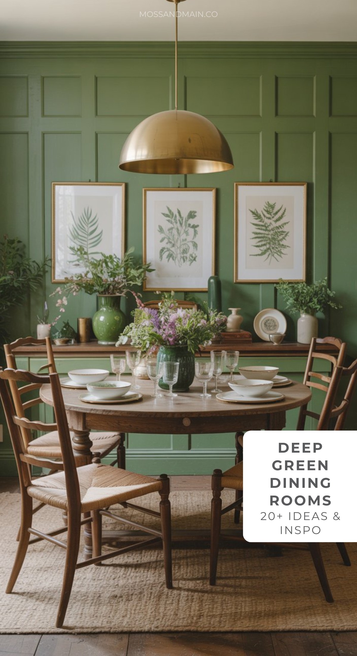 A round wooden dining table set with dishes and flowers sits in a dark green dining room. Wooden chairs surround the table, while botanical prints hang on the paneled wall under a gold pendant light.