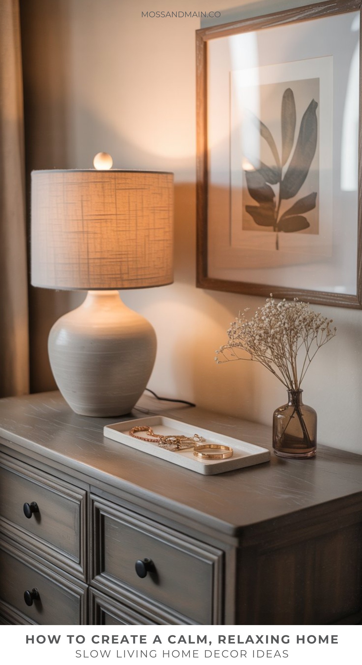 A softly-lit table lamp sits on a wooden dresser next to a framed botanical print and a brown vase with dried flowers—an inviting display that captures the essence of slow living home decor and creates a calm, cozy atmosphere.