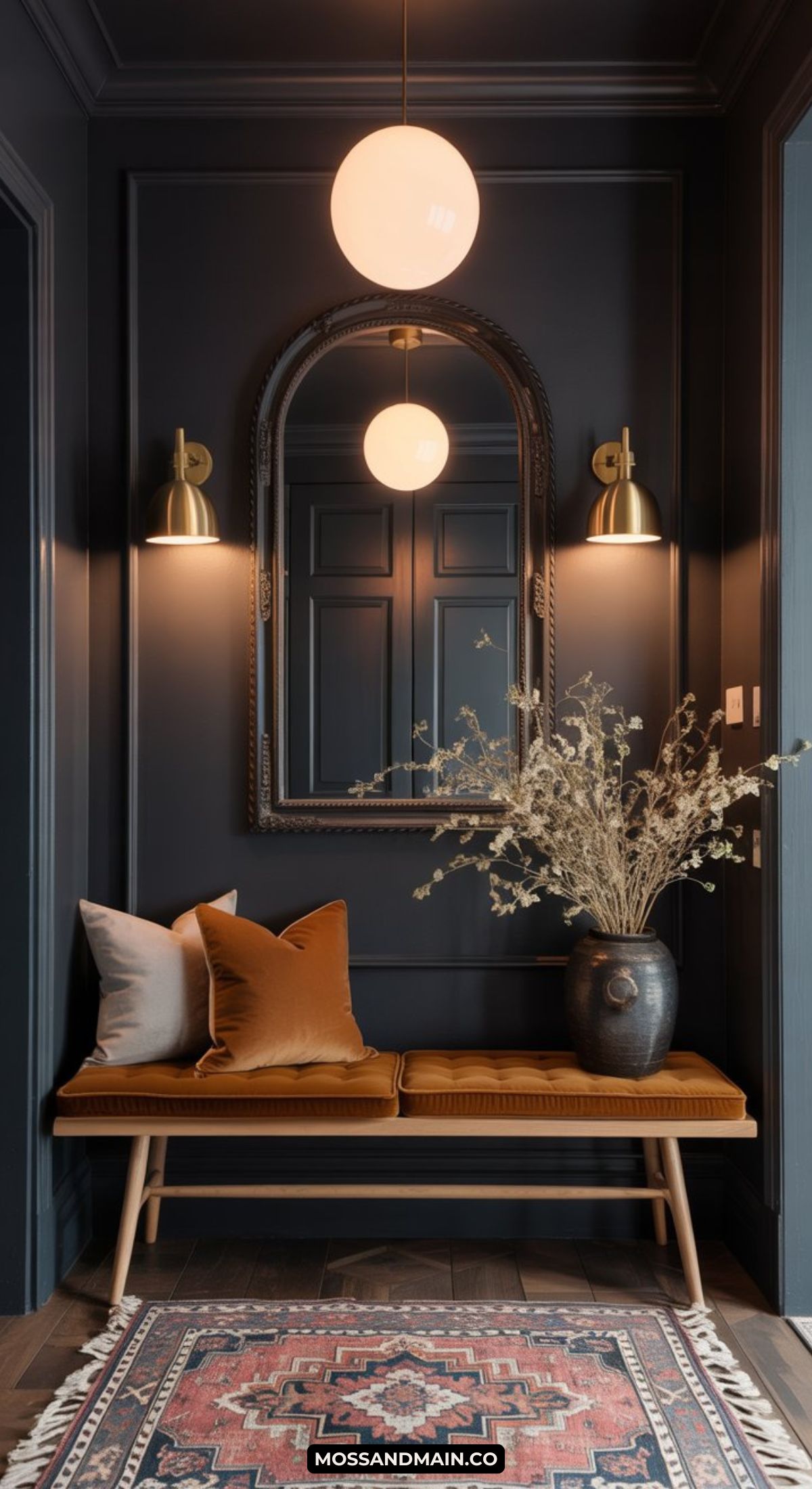 A dark and moody entryway features dark walls, a round mirror, brass sconces, a wooden bench with rust and white cushions, a vase of dried flowers, and a patterned rug. A globe pendant light completes the stylish look.