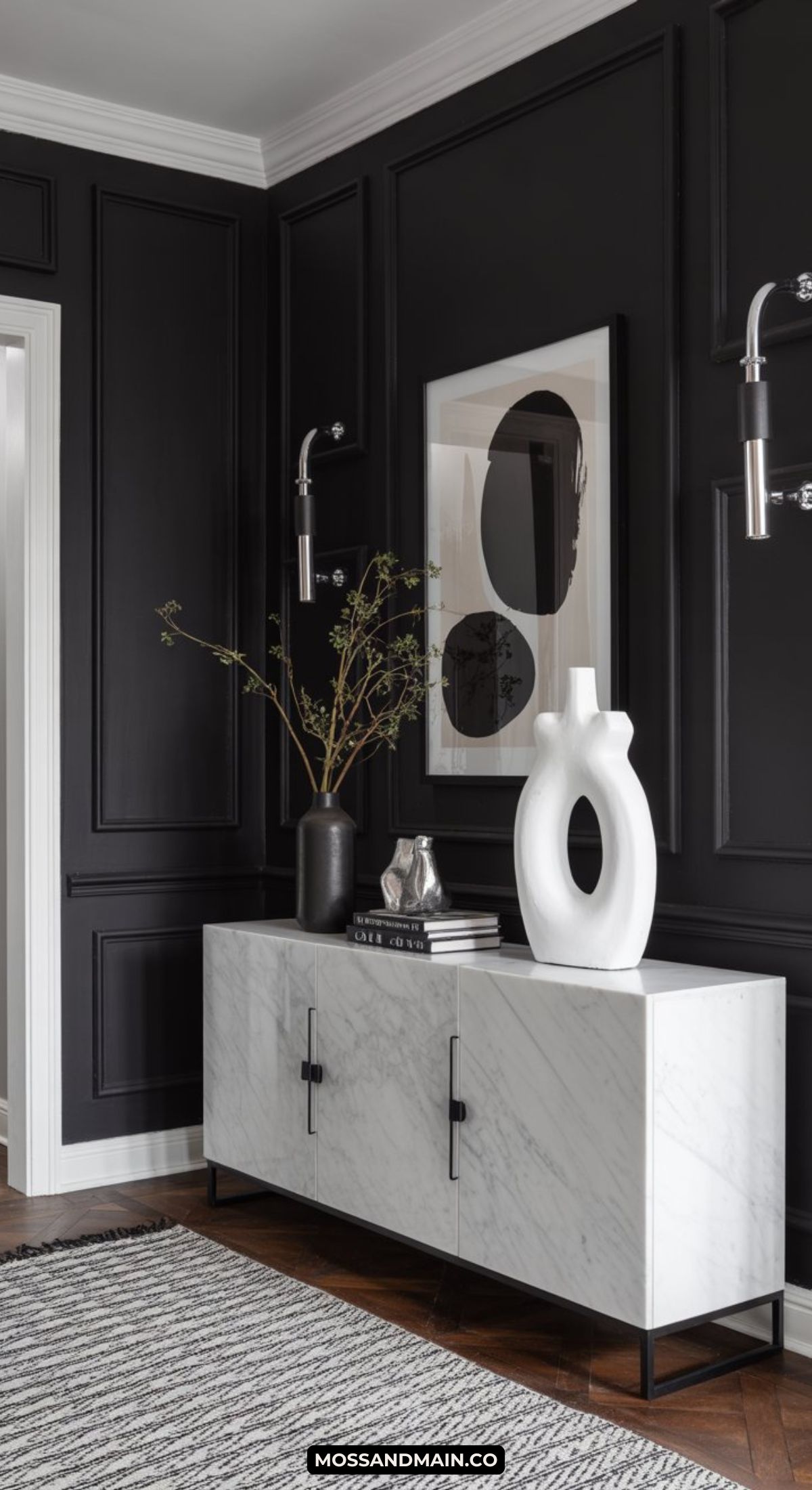 A dark and moody entryway with black paneled walls, a white marble cabinet, minimalist decor including abstract art, a sculptural white vase, a black vase with branches, a silver pitcher, and a striped rug on a wood floor.