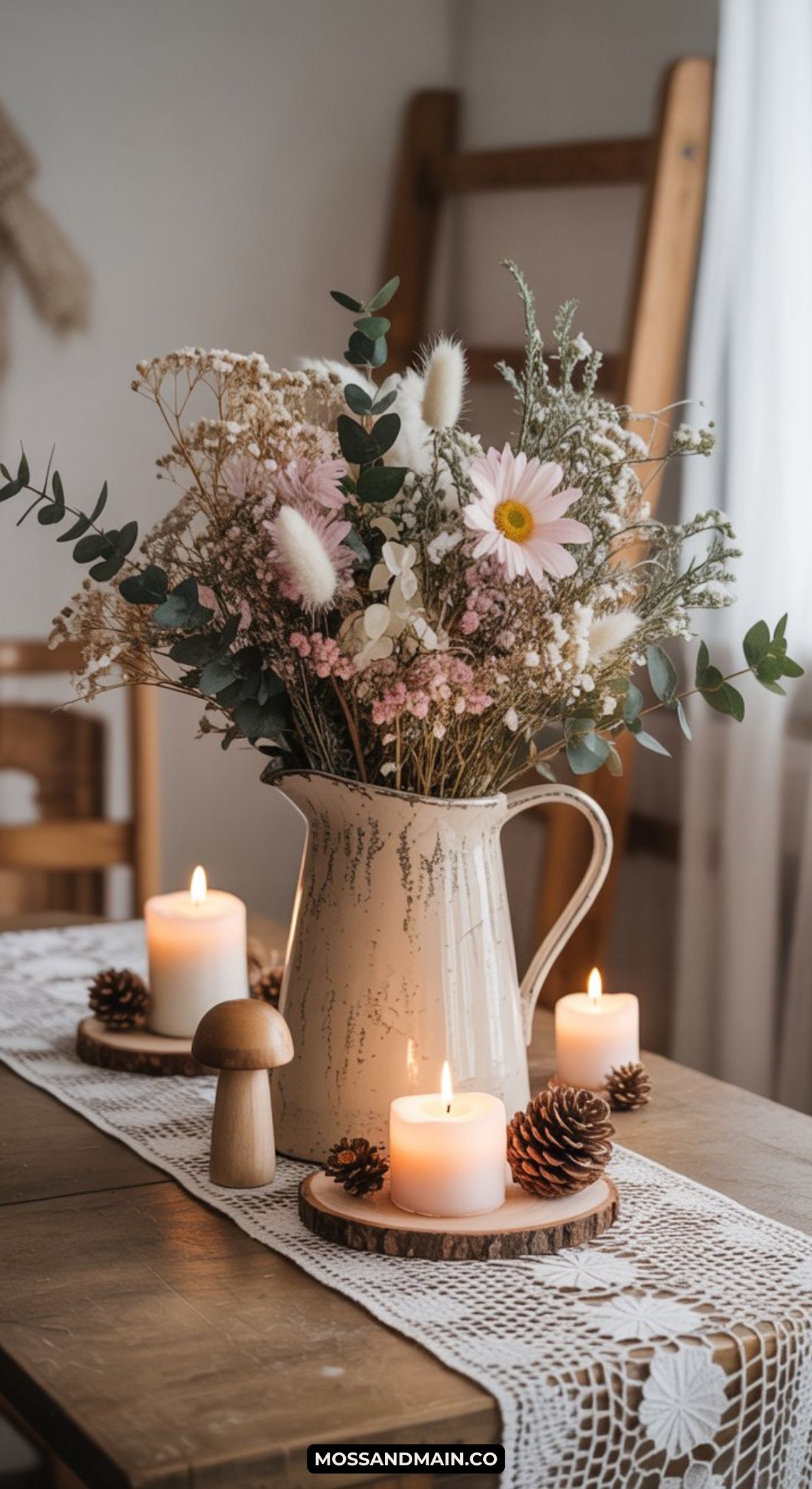 A rustic winter centerpiece features a cream ceramic pitcher filled with wildflowers and greenery, surrounded by lit candles, pinecones, and a wooden mushroom on a lace table runner in a cozy room.