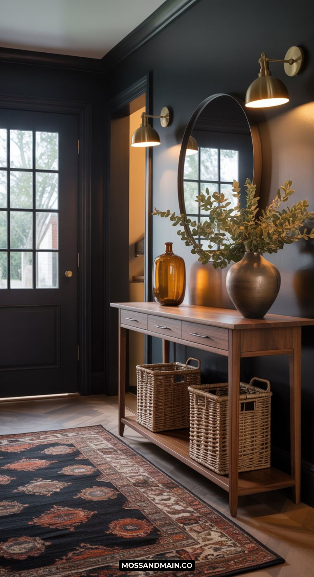 A dark and moody entryway features a black door, dark walls, a wooden console table with baskets, a round mirror, gold wall sconces, a patterned rug, and decorative vases filled with greenery.