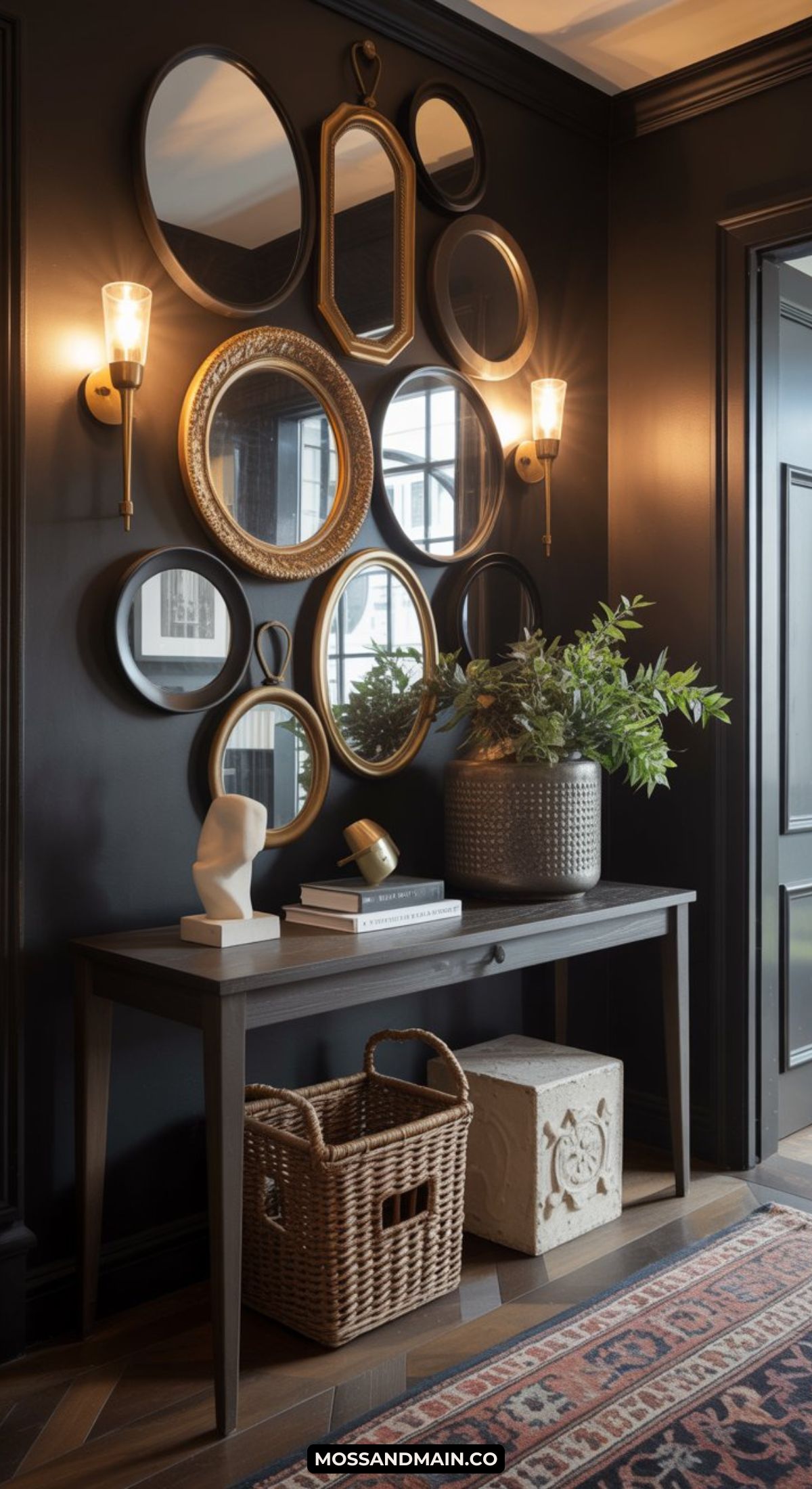 A dark and moody entryway features black walls, a console table with books, a white sculpture, a woven basket, and a potted plant. Multiple gold-framed mirrors are arranged above in an elegant gallery style.