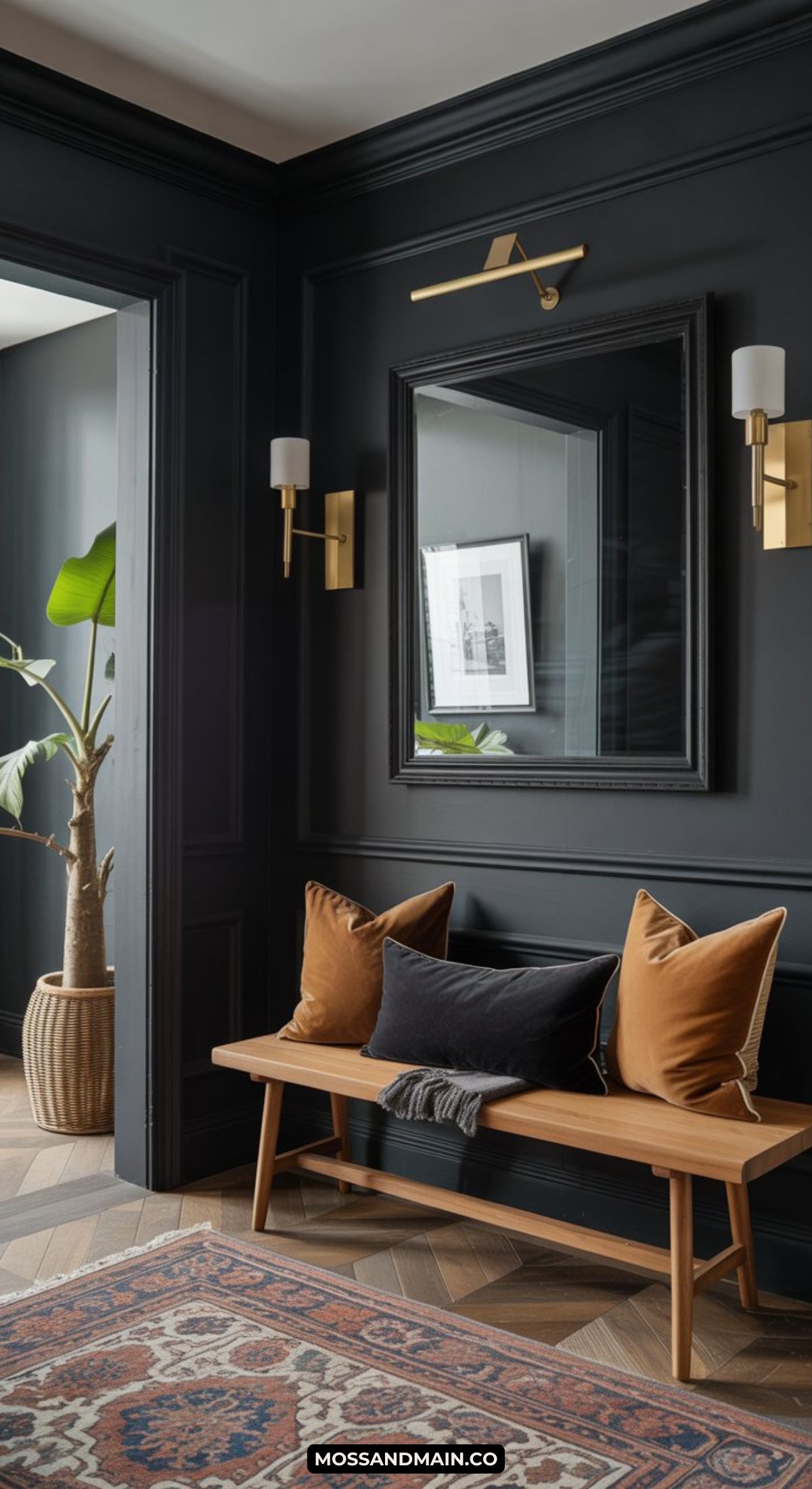 A dark and moody entryway features dramatic walls, a large mirror, gold wall sconces, a wooden bench with black and brown pillows, a patterned rug, and a potted plant in the corner.