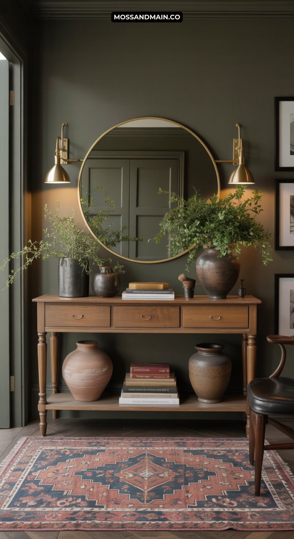 A wooden console table with drawers, decorated with ceramic vases, greenery, and books, sits below a round gold mirror and two wall sconces. A patterned rug and framed wall art enhance this dark and moody entryway’s cozy, modern vibe.