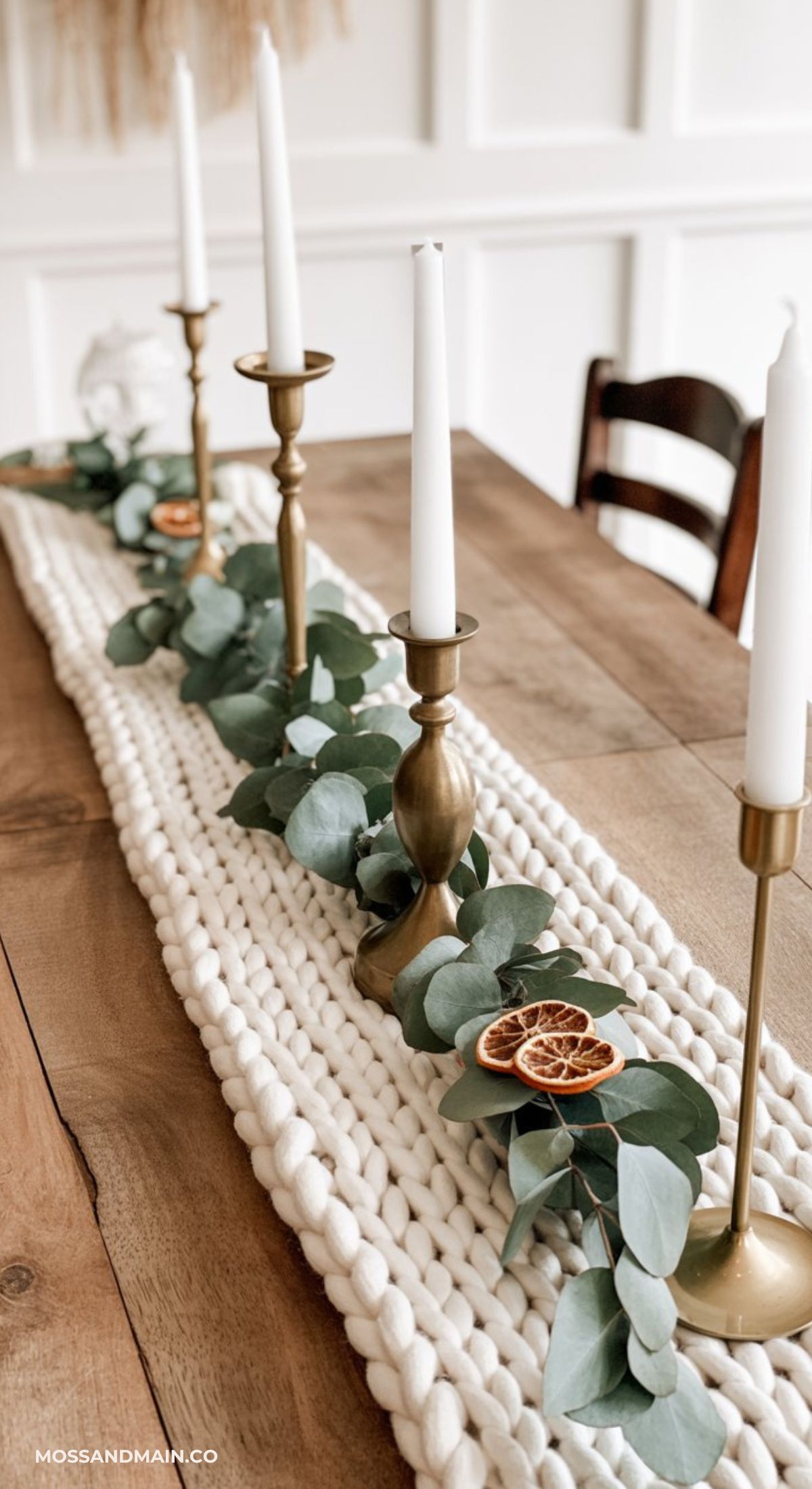 A wooden dining table decorated with a chunky knit runner, greenery, dried orange slices, and brass candle holders with white taper candles in a row—perfect for inspiring cozy winter centerpiece ideas.