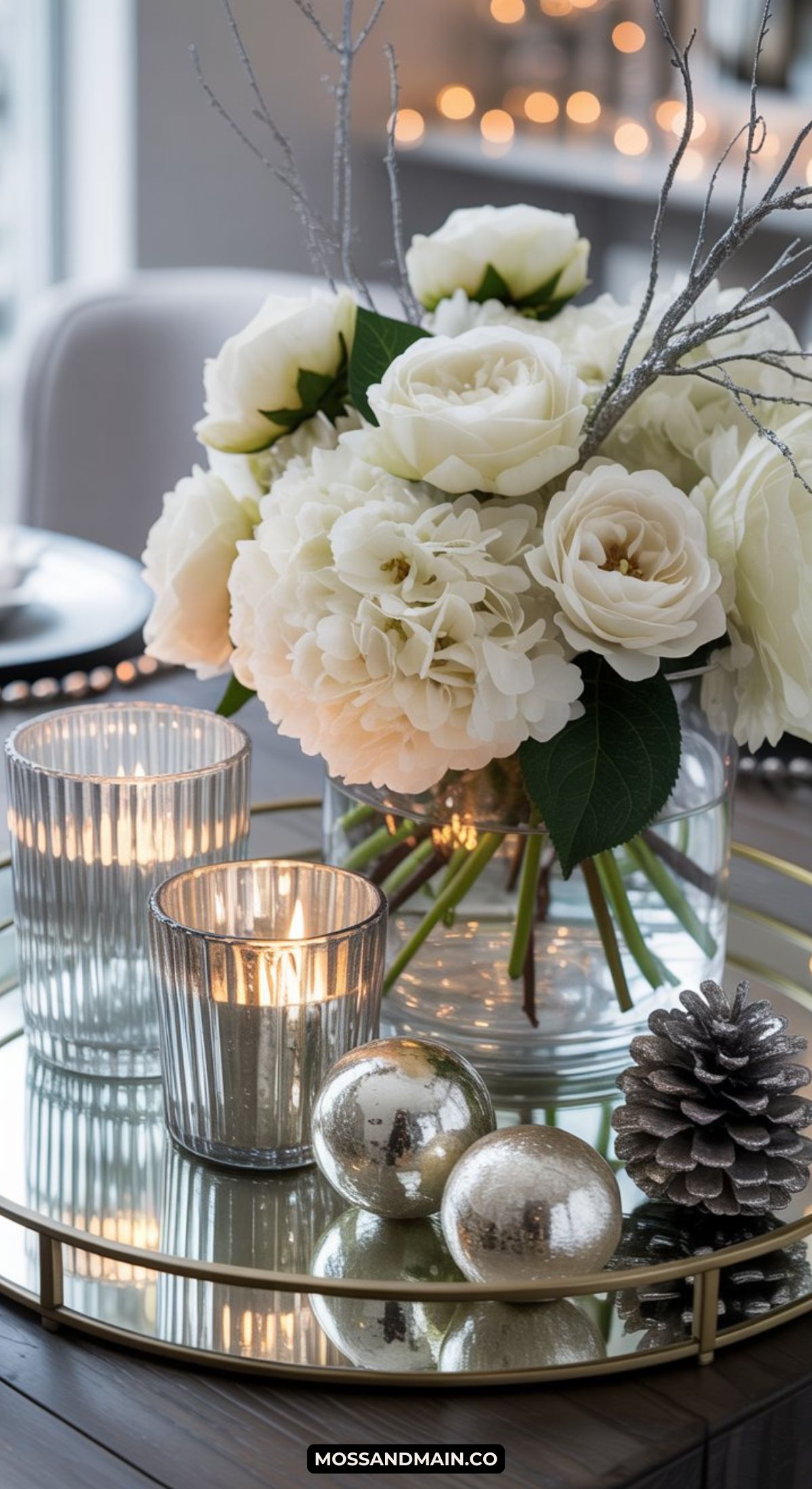 A glass tray holds a bouquet of white flowers, two lit candles in ribbed glass holders, silver ornaments, and a pine cone, creating an elegant and cozy Winter Centerpiece for your table.