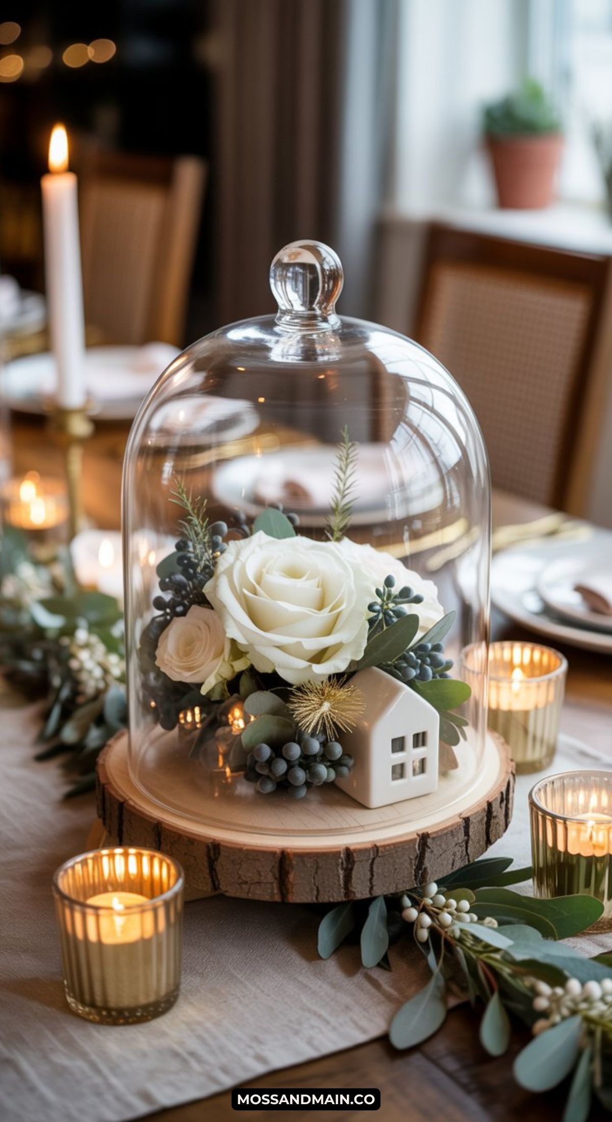 A glass cloche covers white roses, greenery, berries, and a small decorative house on a wooden base—perfect inspiration for Winter Table Decor—surrounded by candles and foliage on an elegant dining table.