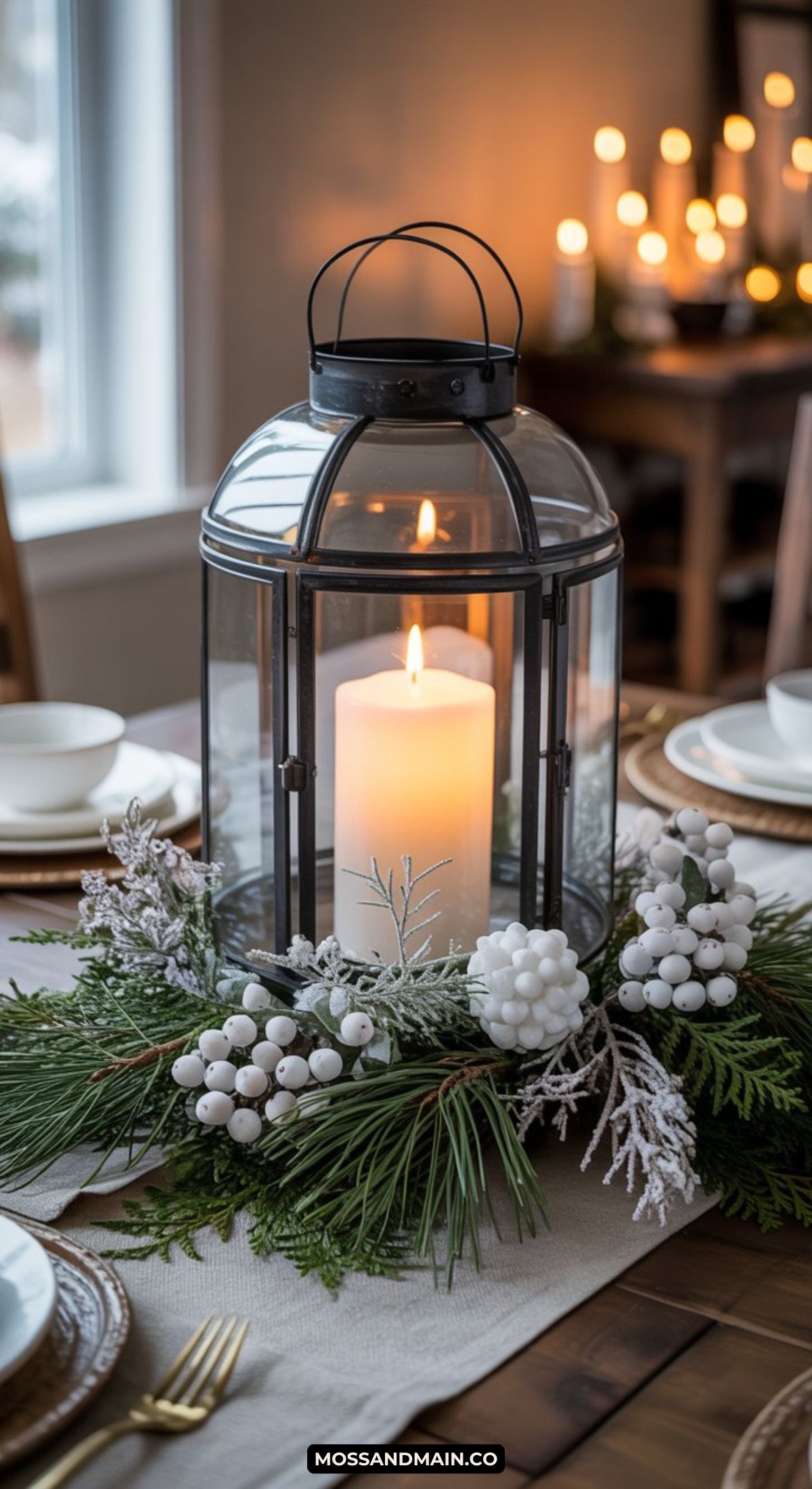 A large glass lantern with a lit white candle sits on a dining table, surrounded by pine branches, white berries, and festive greenery—an elegant example of Winter Centerpiece Ideas. More candles glow in the background, enhancing the cozy winter table decor.