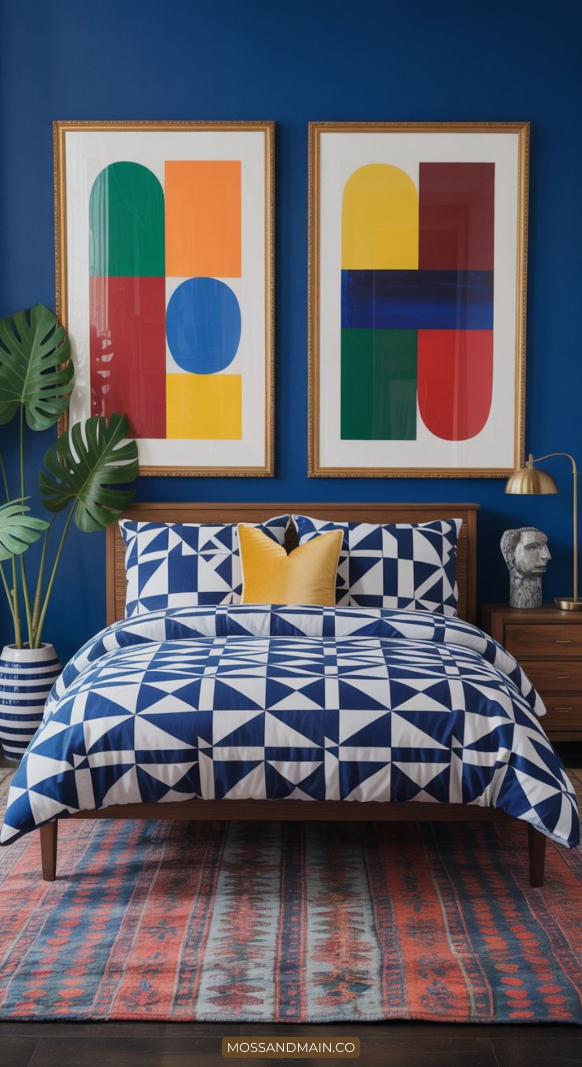 A modern bedroom featuring maximalist color with a blue geometric bedspread, yellow accent pillow, and patterned rug. Two bold abstract art prints hang on a deep blue wall, while a plant and gold desk lamp decorate the bedside table.
