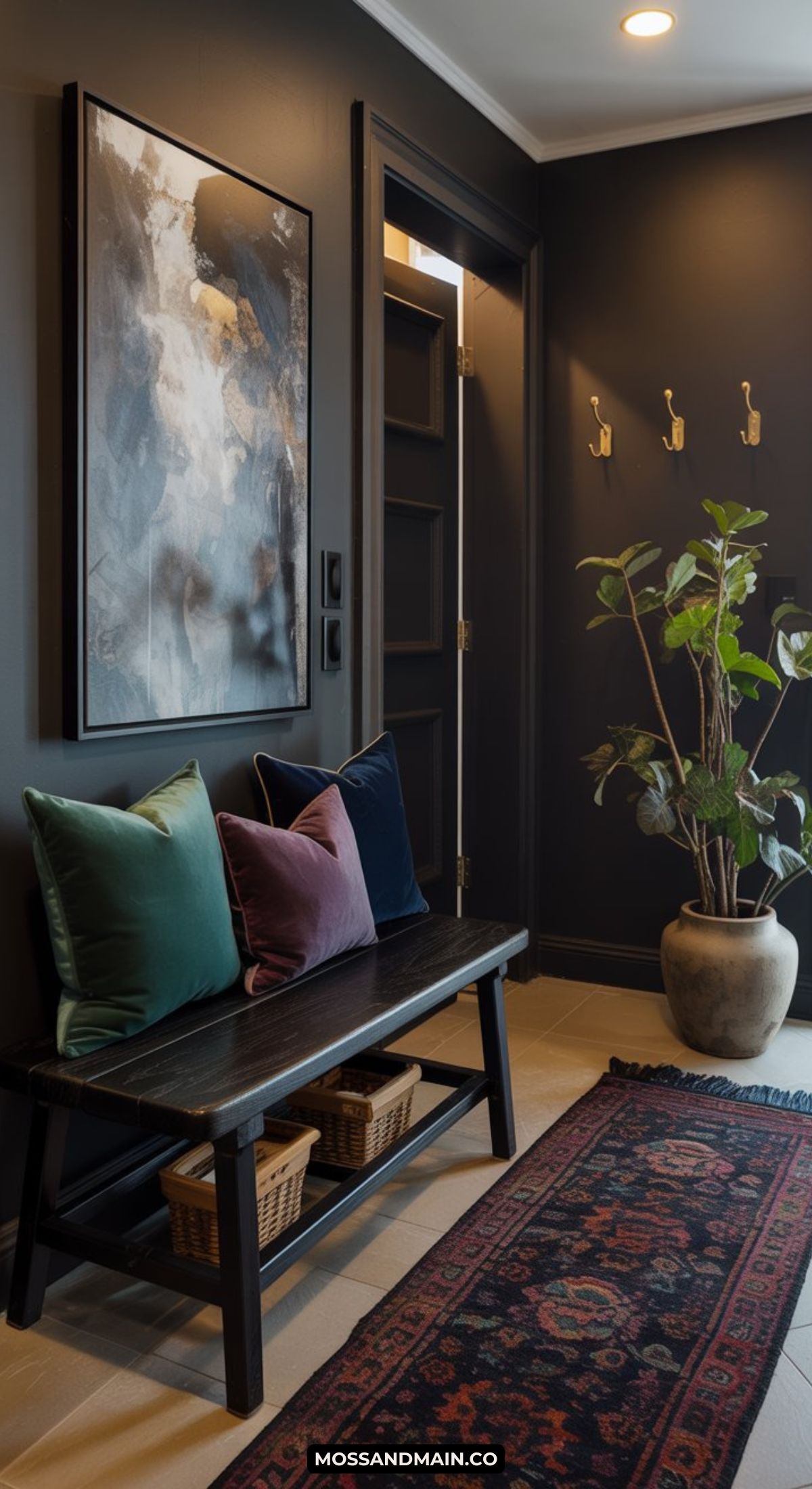 A dark and moody entryway features dark walls, a wooden bench with green, blue, and purple pillows, baskets underneath, a large abstract painting, a potted plant, wall hooks, and a patterned rug on the floor.