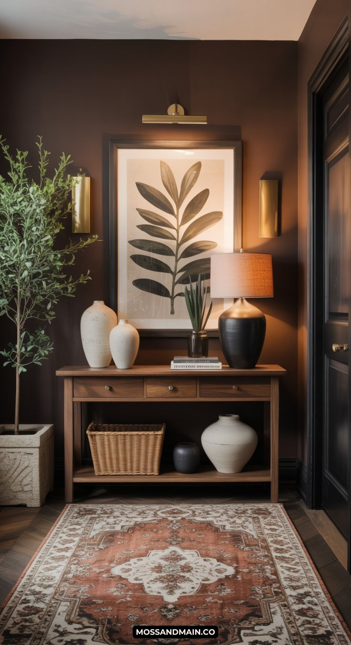 A warm, stylish entryway features a wooden console table with vases, a basket, a lamp, and a framed botanical print. A hint of dark and moody entryway style comes through in the soft sconce lighting and richly patterned rug.
