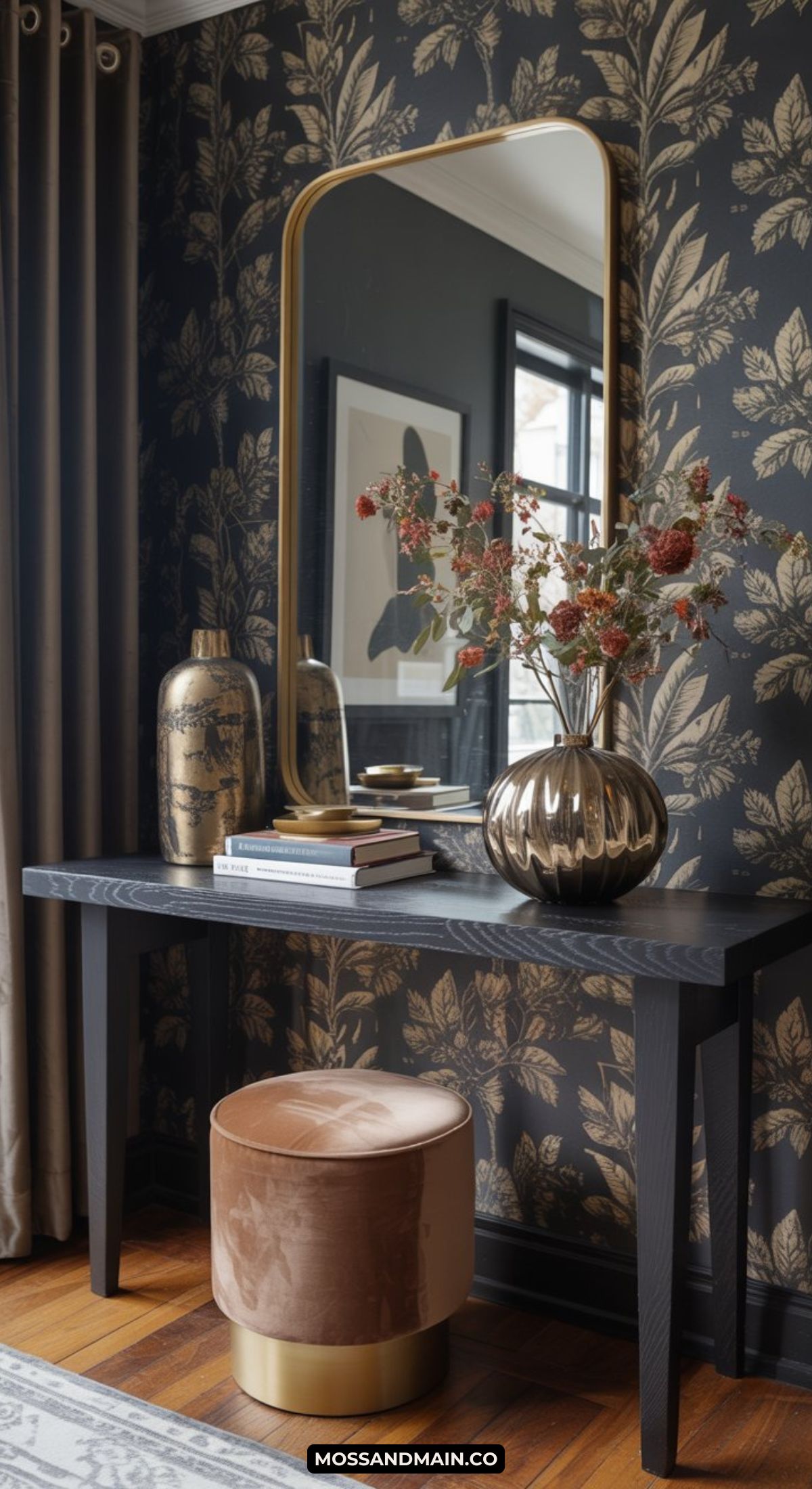 A stylish dark and moody entryway features a black console table with gold decor, books, and a vase of dried flowers, set against dark floral wallpaper, a large framed mirror, a beige velvet stool, and a nearby curtained window.