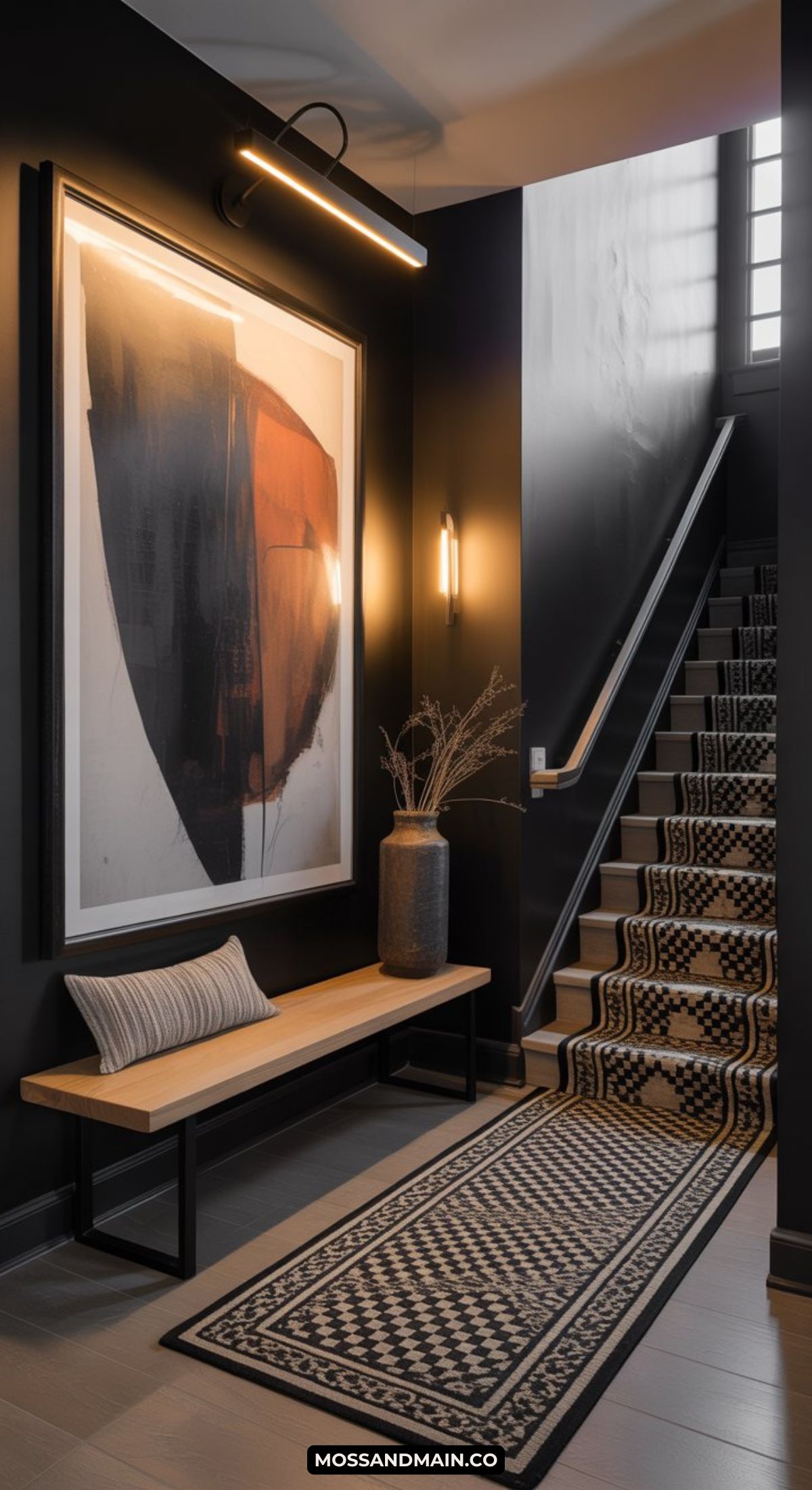 A modern, dark and moody entryway features dark walls, a large abstract artwork, a wooden bench with a striped pillow, a patterned rug, dried branches in a vase, and a staircase with a matching runner carpet.