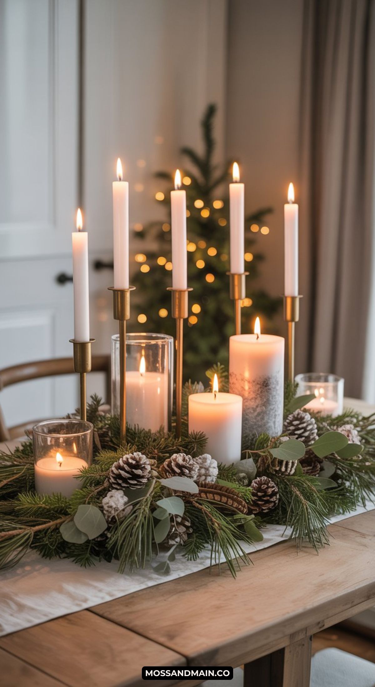 A festive winter centerpiece with lit white candles of varying heights, surrounded by pine branches, pine cones, eucalyptus leaves, and glass candle holders on a wooden table. A blurred Christmas tree glows softly in the background.