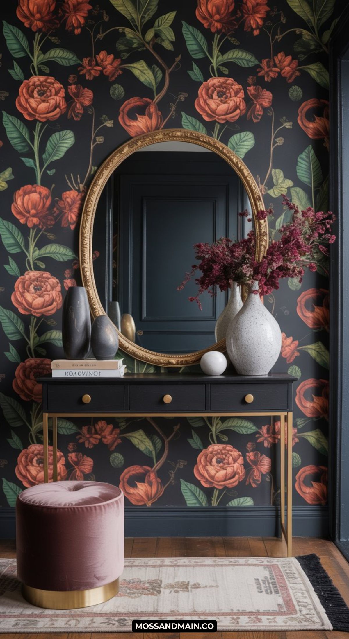 A stylish, dark and moody entryway with bold floral wallpaper, a round gold-framed mirror, a black and gold console table, marble vases with red flowers, stacked books, and a pink velvet ottoman.