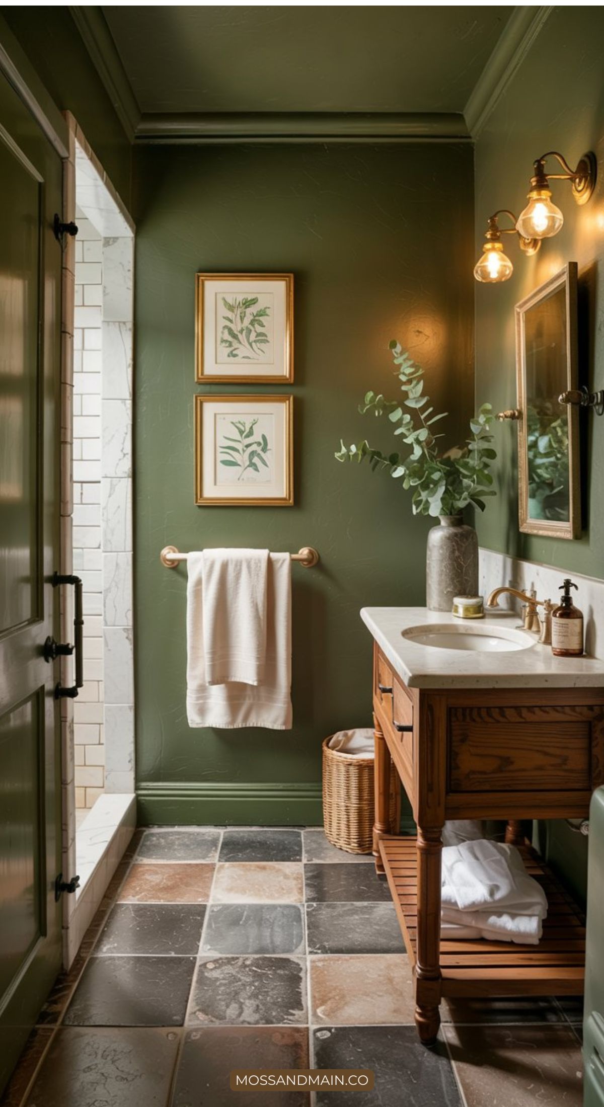 A bathroom with dark green walls, a wooden vanity with a white countertop, and trending bathroom elements like a round vase with greenery. Two botanical prints, a towel rack, tiled floor, and shower showcase fresh 2026 Bathroom Trends in design.