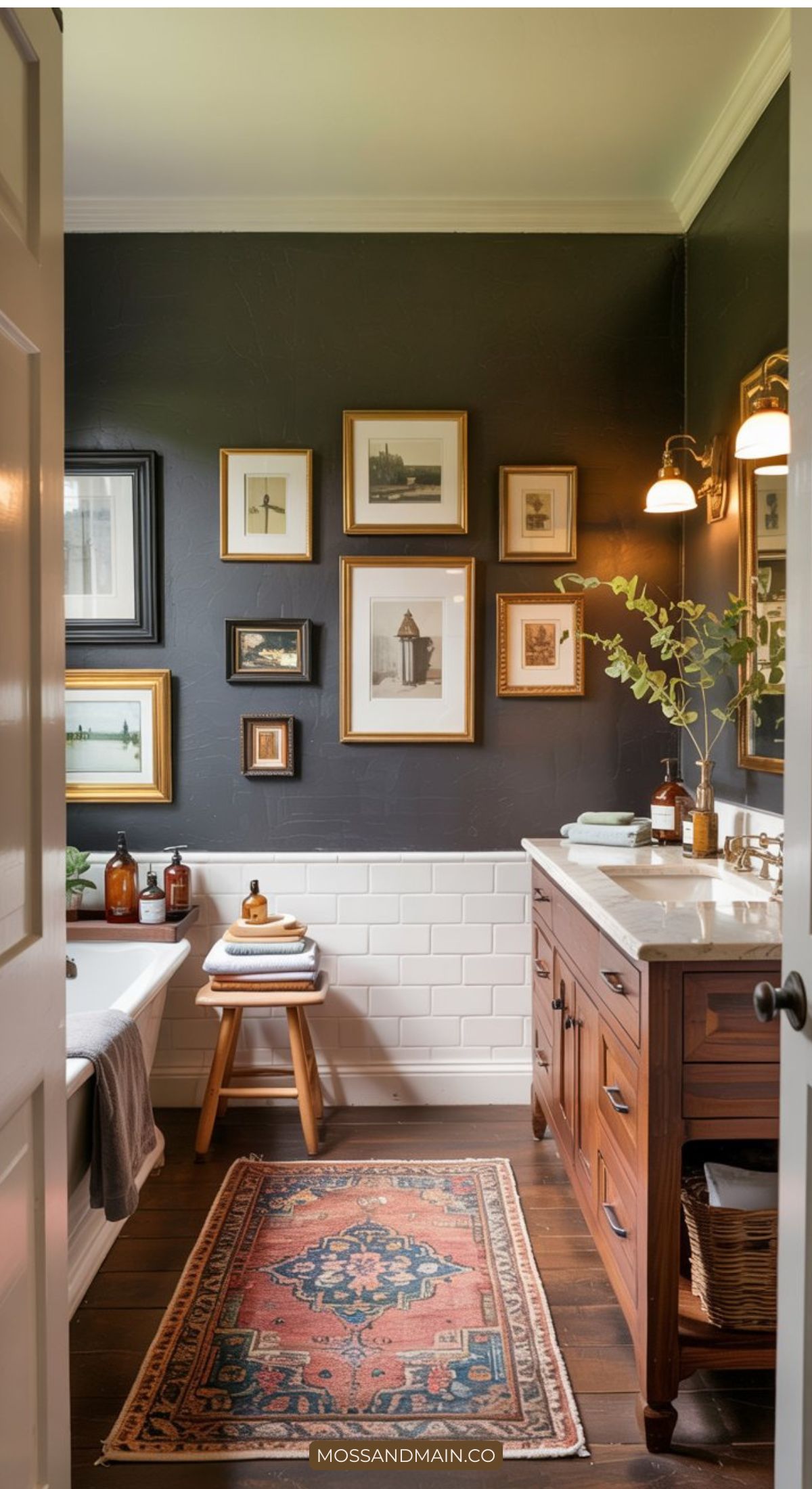 A cozy bathroom design features dark walls with framed art, a freestanding bathtub, and wooden vanity—highlighting 2026 bathroom trends like brass fixtures, potted greenery, and white subway tile wainscoting. Natural light brightens the space.