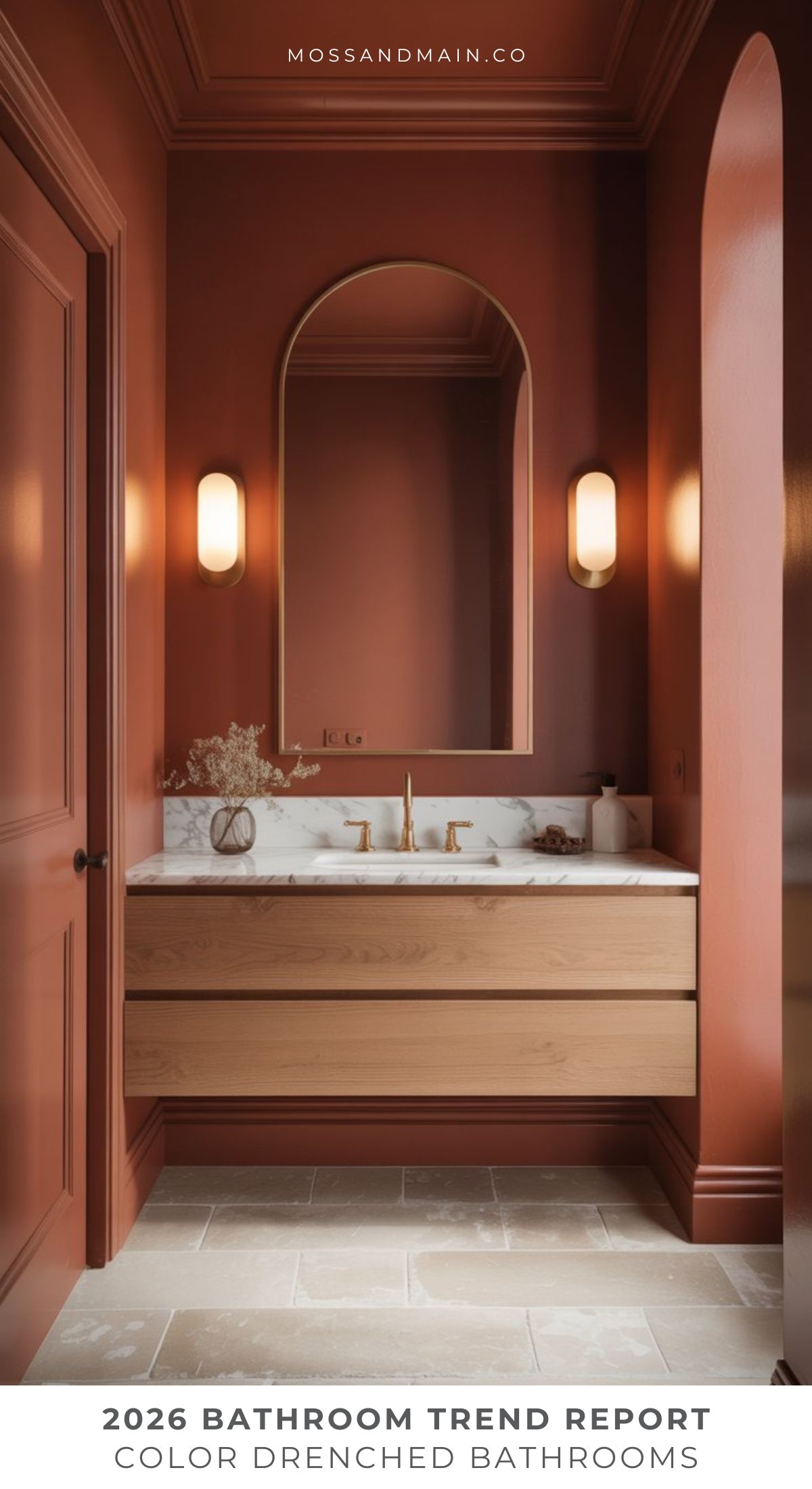 A modern bathroom with terracotta-colored walls, a wooden vanity with a white countertop, brass fixtures, a large oval mirror, wall sconces, and a small vase with flowers. Text reads: 2026 Bathroom Trend Report: Color Drenched Bathroom Design Trends.
