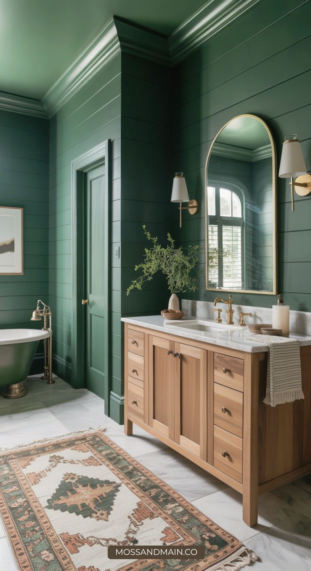 A stylish bathroom embracing bathroom design trends with dark green shiplap walls, a wooden vanity and marble countertop, a round mirror, sconces, a green bathtub, a window, and a patterned rug on a marble floor.