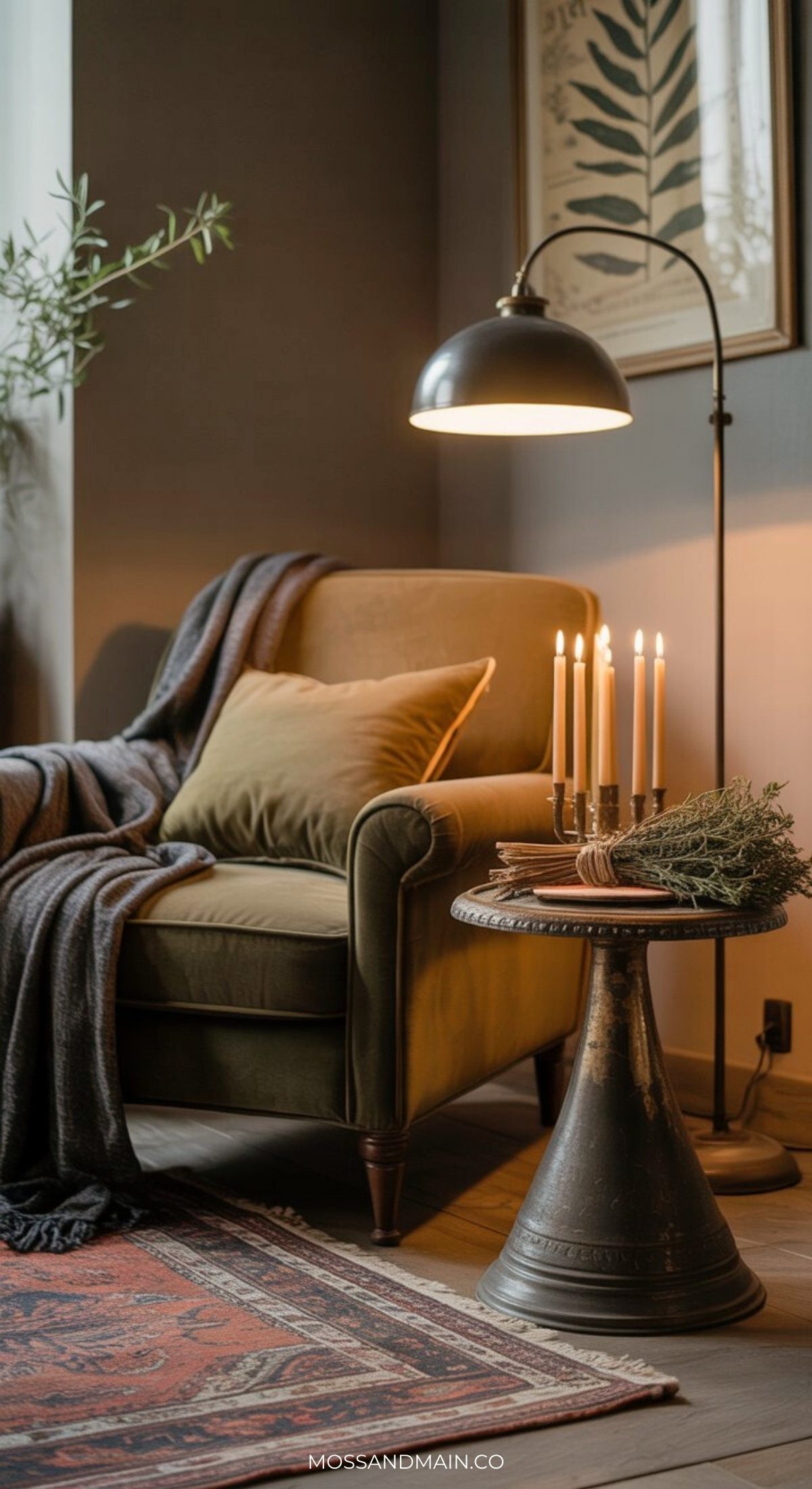 A cozy reading nook with an olive green armchair draped in a dark blanket, a side table holding lit candles, books, and greenery—all softly lit by a black floor lamp—creates an earthy living room with a natural aesthetic. A botanical print decorates the wall.