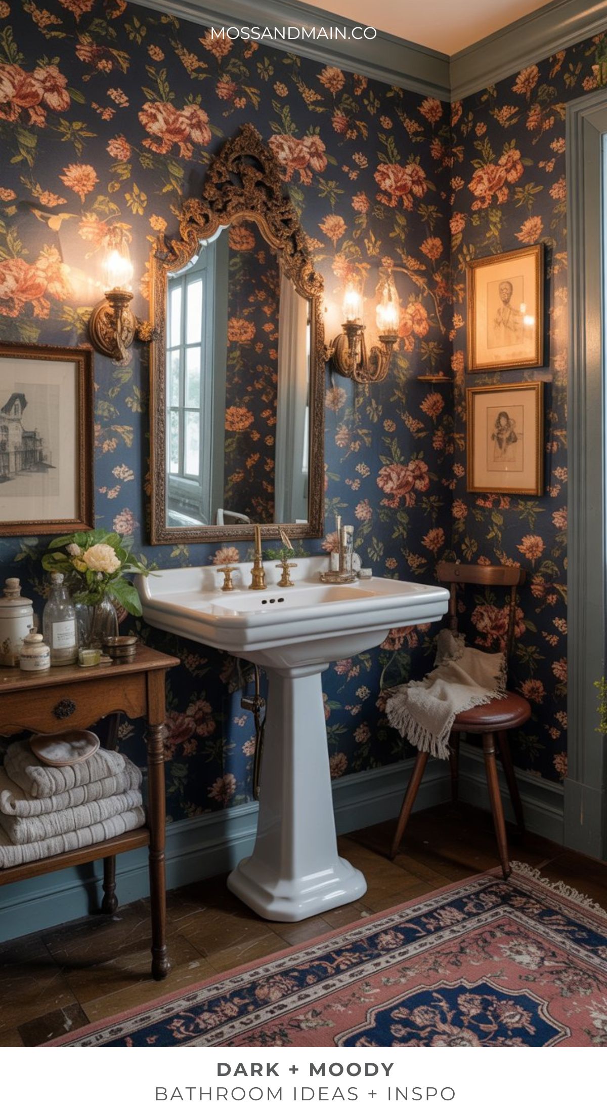 A dramatic bathroom featuring vintage-style dark floral wallpaper, ornate mirrors, moody wall sconces, a white pedestal sink, framed art, a wooden chair with a towel, and decorative touches on the shelf and rug.
