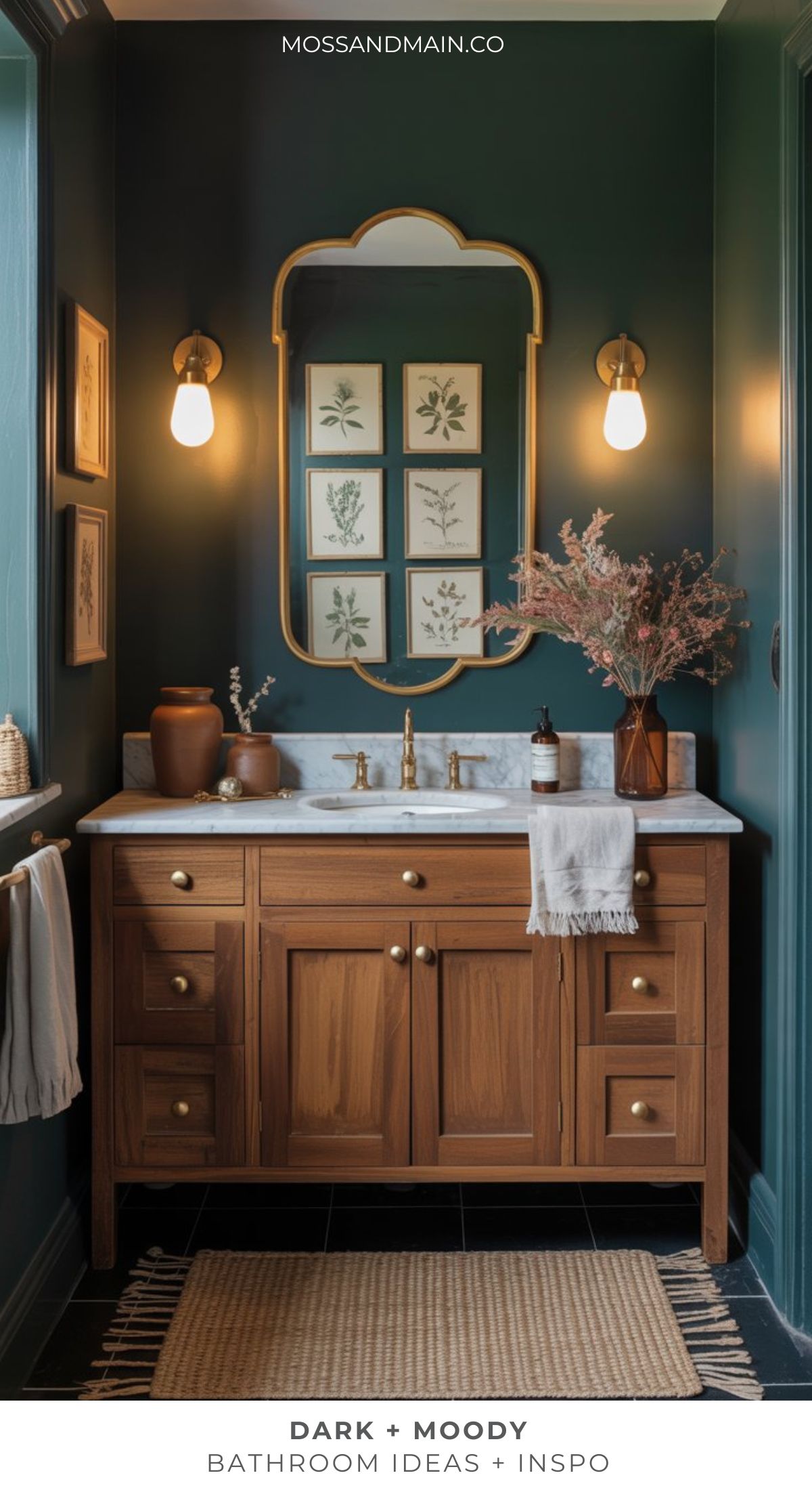 A bathroom with dark green walls, a wooden vanity with brass hardware, a marble countertop, and botanical wall art embodies moody bathroom ideas. Warm lighting and dried flowers complete this dramatic bathroom look. Text reads Dark + Moody Bathroom Ideas + Inspo.