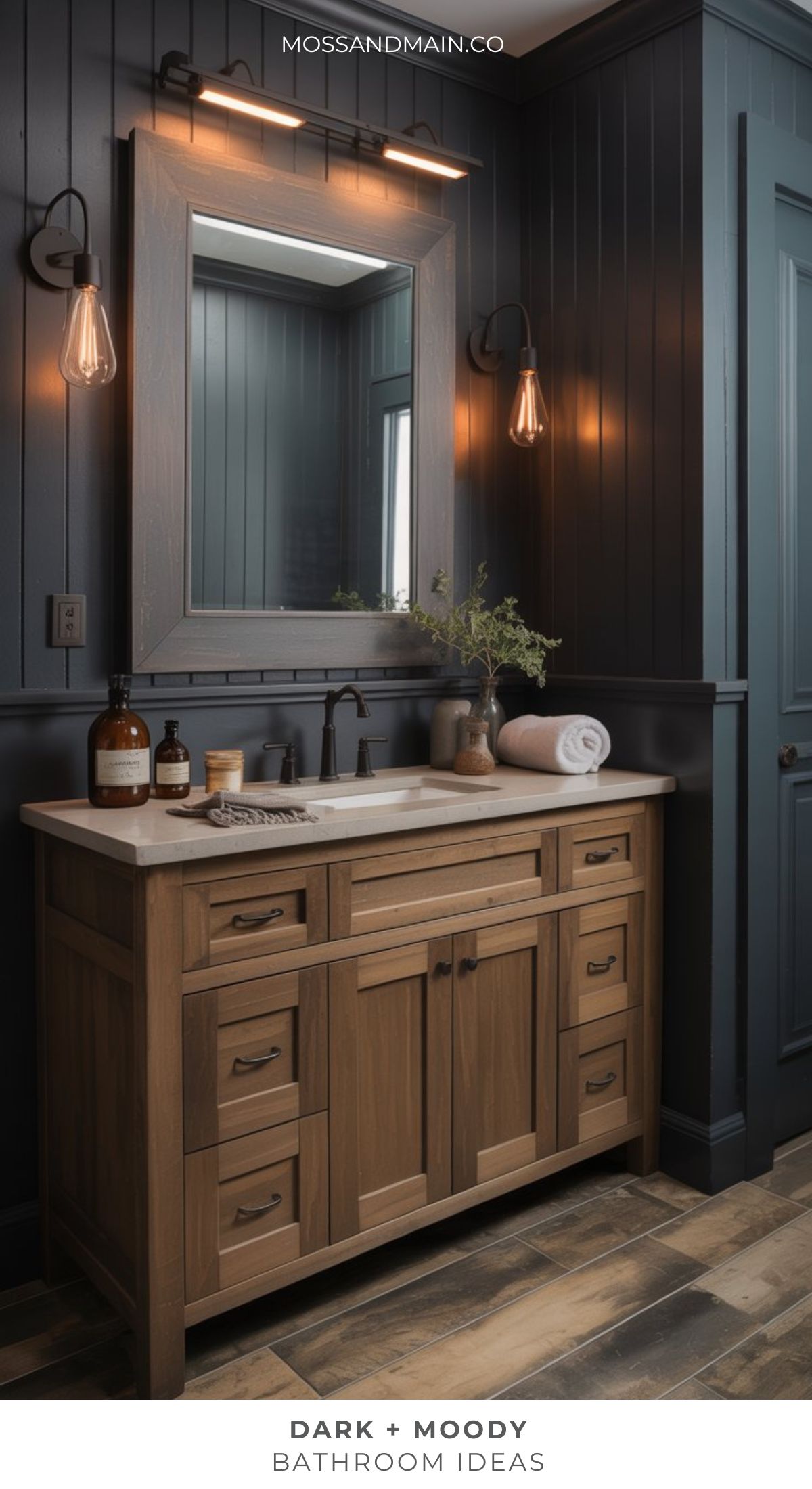 A dramatic bathroom with dark paneled walls, a wooden vanity topped with white, modern sconces, and a large framed mirror creates a sophisticated, moody bathroom atmosphere.