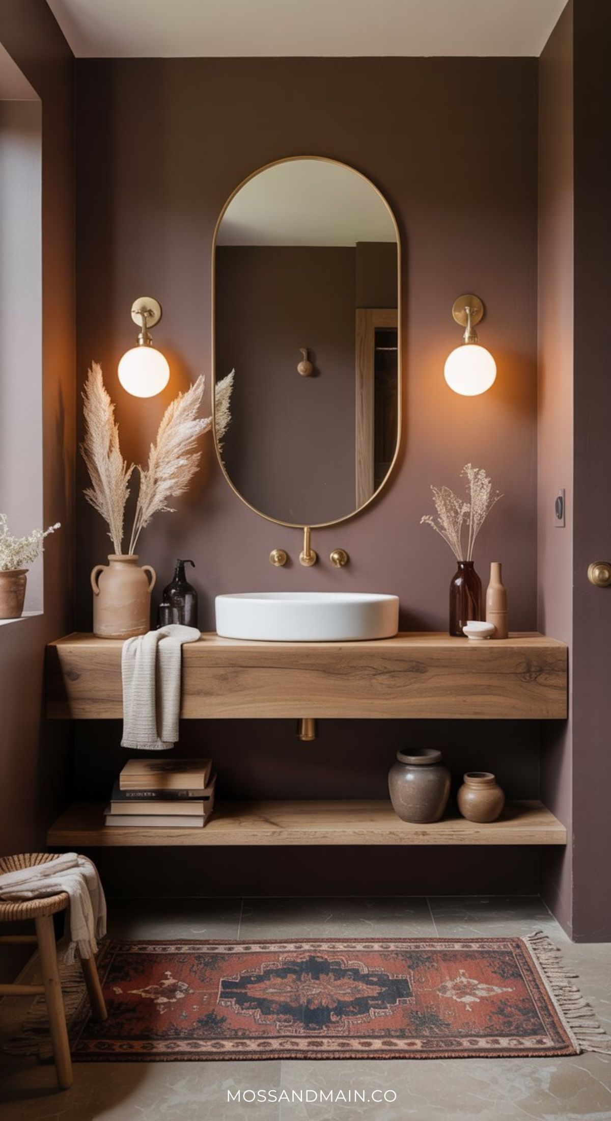 A modern, dramatic bathroom with brown walls, a wooden floating vanity, oval mirror, brass fixtures, globe wall lights, pampas grass in vases, ceramic decor, and a patterned rug—perfect for moody bathroom ideas.