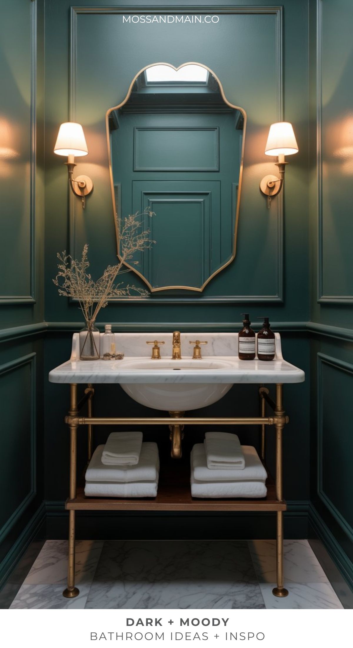 A stylish, dramatic bathroom with dark green paneled walls, a gold-framed mirror, wall sconces, a white sink with gold fixtures, and neatly folded towels on a wooden shelf. Bottles and a vase decorate the countertop—perfect for moody bathroom ideas.