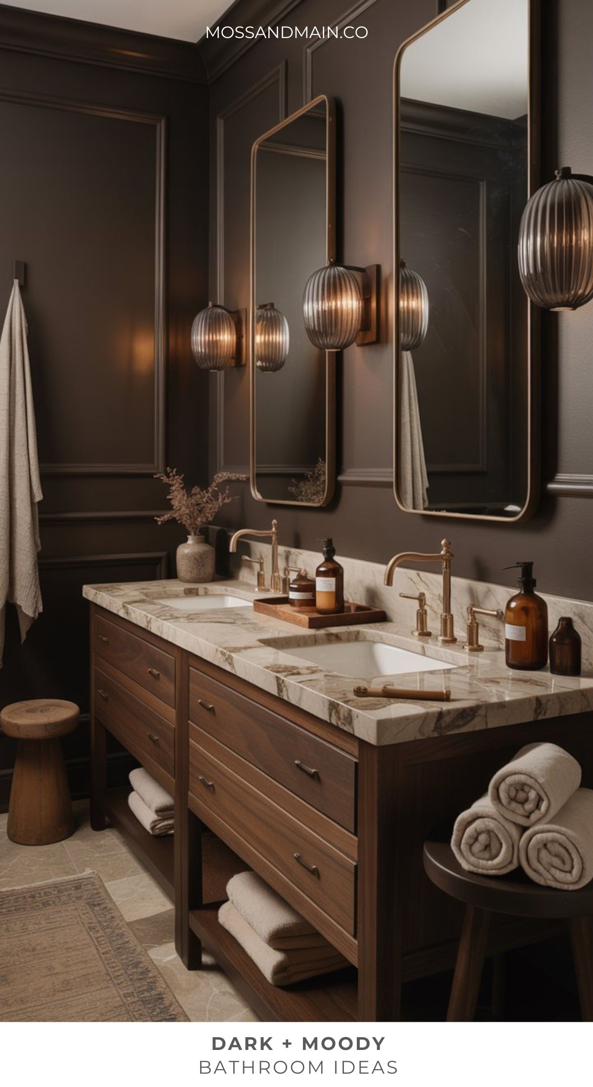 A stylish, dark moody bathroom features a double marble sink vanity, dark wood cabinets, bronze-framed mirrors, warm sconces, and neatly rolled towels that create a dramatic atmosphere.