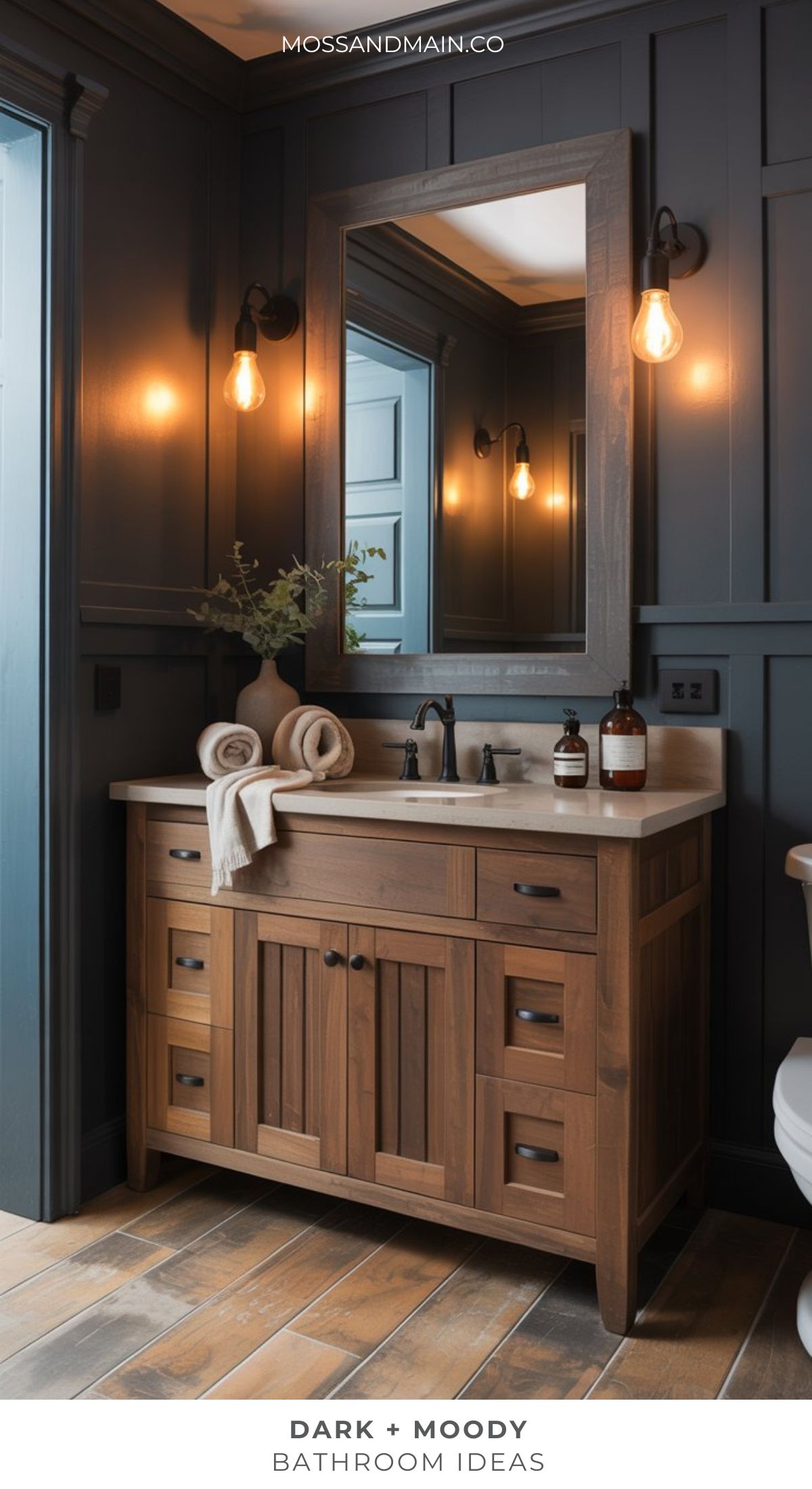 A stylish moody bathroom with dark blue paneled walls, a wooden vanity with a beige countertop, black faucet, rolled towels, decorative vases, and exposed bulb wall sconces creates a warm, atmospheric retreat—perfect for dark bathroom ideas.