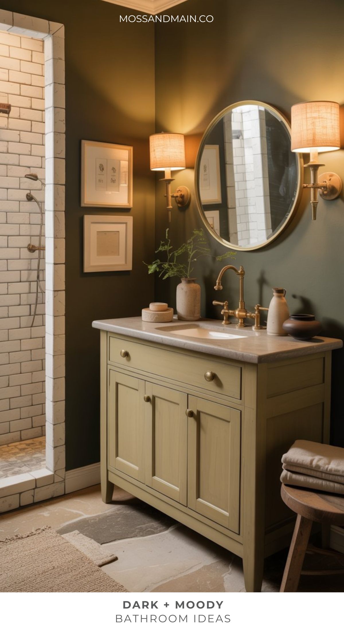 A dark moody bathroom with a deep green wall, round mirror, gold fixtures, and beige vanity with marble countertop. Dramatic bathroom touches include framed art, wall sconces, and a peek of the cozy adjacent tiled shower.