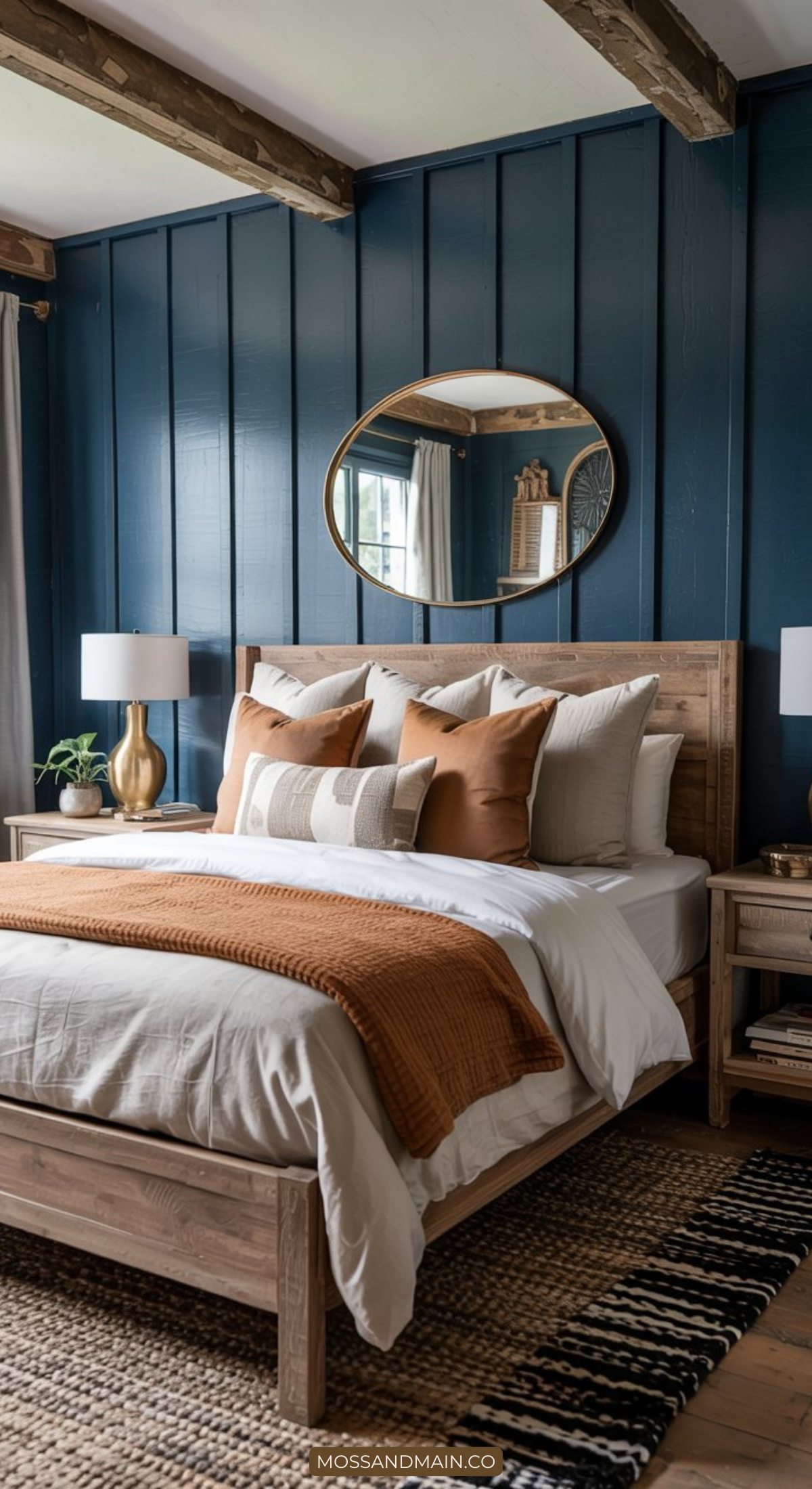 A cozy bedroom showcases modern farmhouse style with a wooden bed, white and tan bedding, a rust-colored throw, and plush pillows. A round mirror hangs above the bed on a navy-paneled wall, flanked by matching lamps on bedside tables.