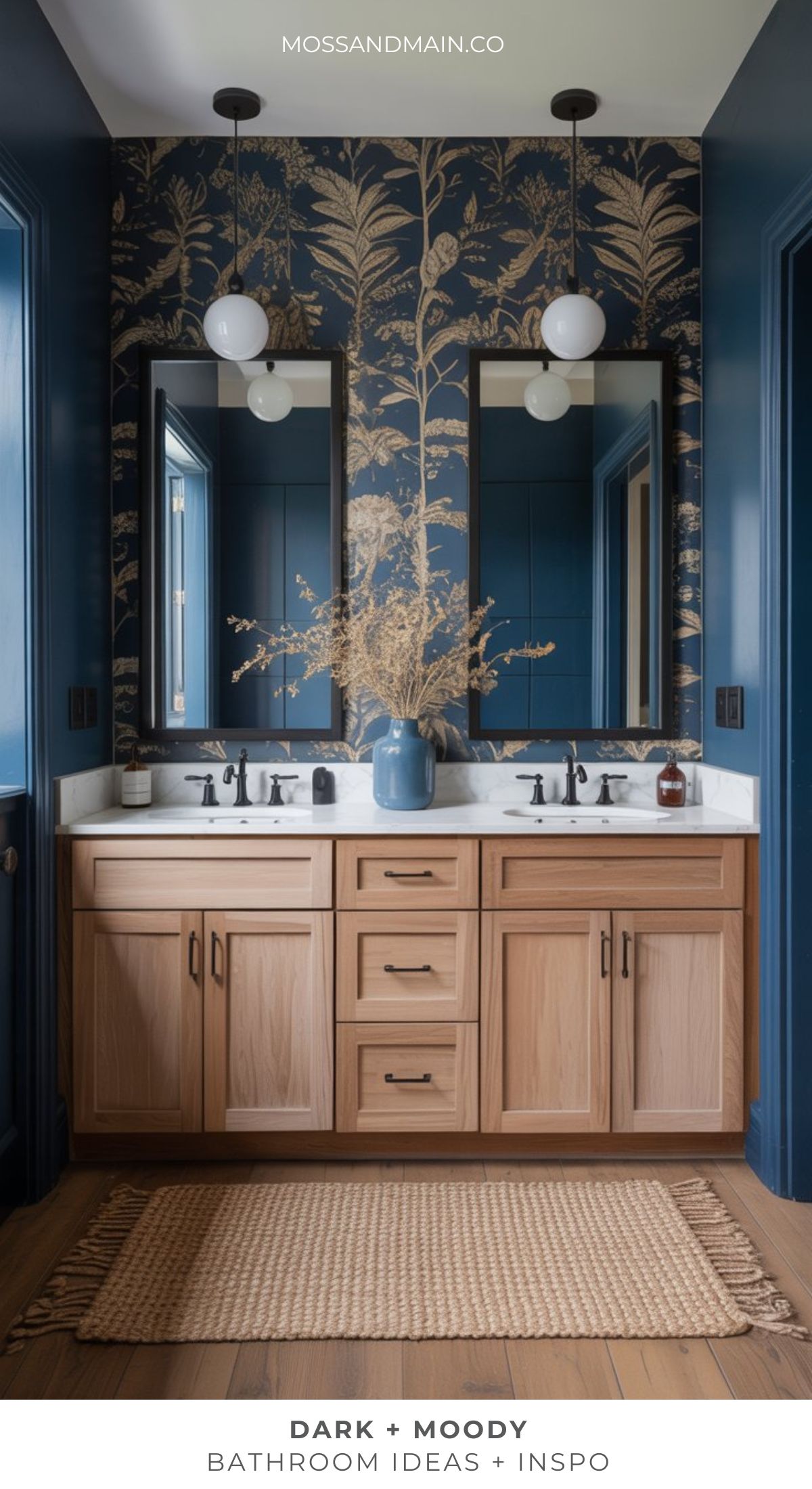 A moody bathroom with double sinks, light wood cabinets, and dark faucets. Deep blue walls feature gold botanical wallpaper, while a woven rug warms the wood floor. Two mirrors and globe lights complete this dramatic bathroom retreat.