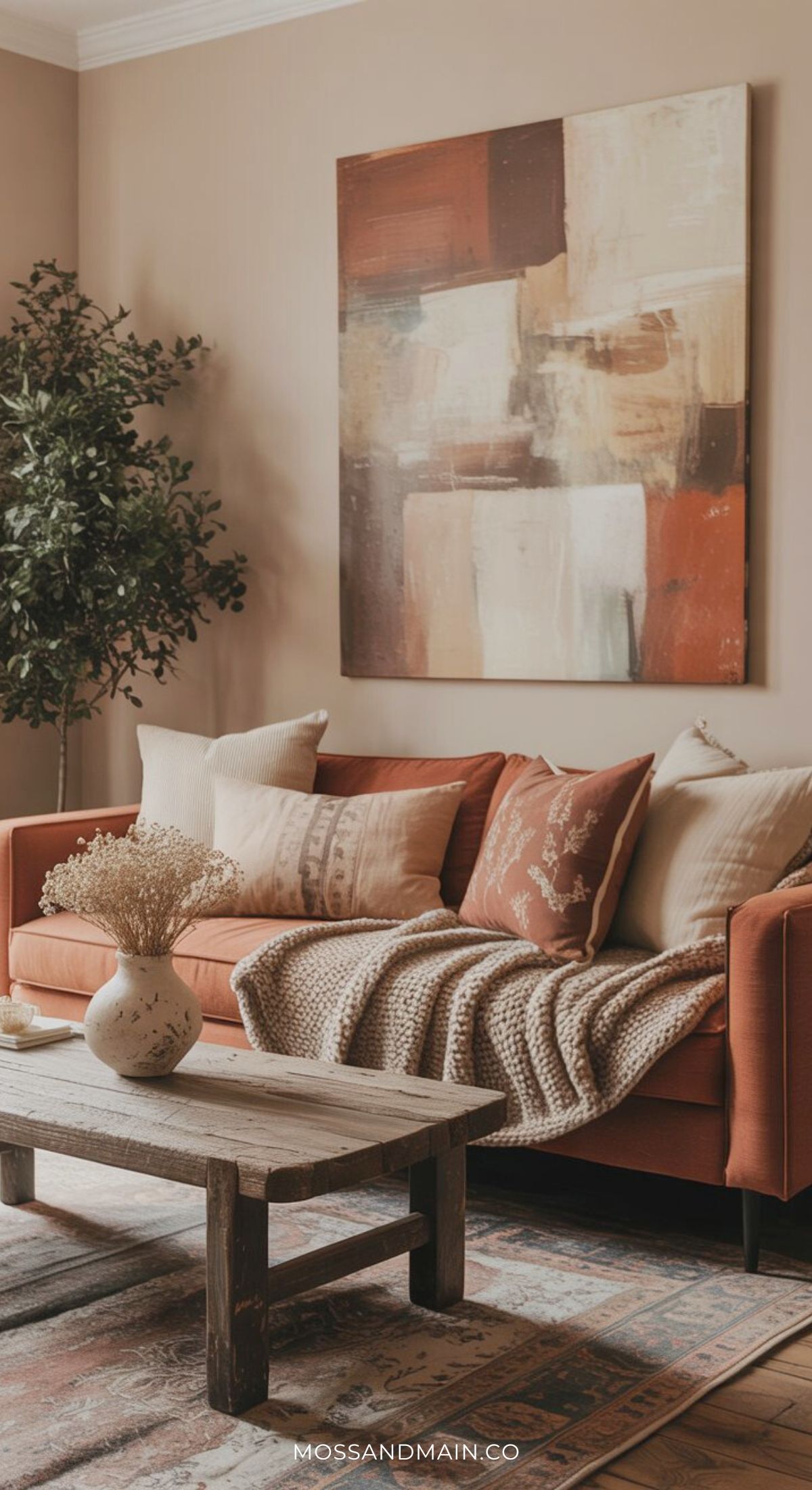 A cozy living room with a rust couch, assorted neutral pillows, a knitted throw, wooden coffee table with a ceramic vase of dried flowers, large abstract painting, and tall green plant—perfect inspiration for fresh living room ideas beyond boring spaces.