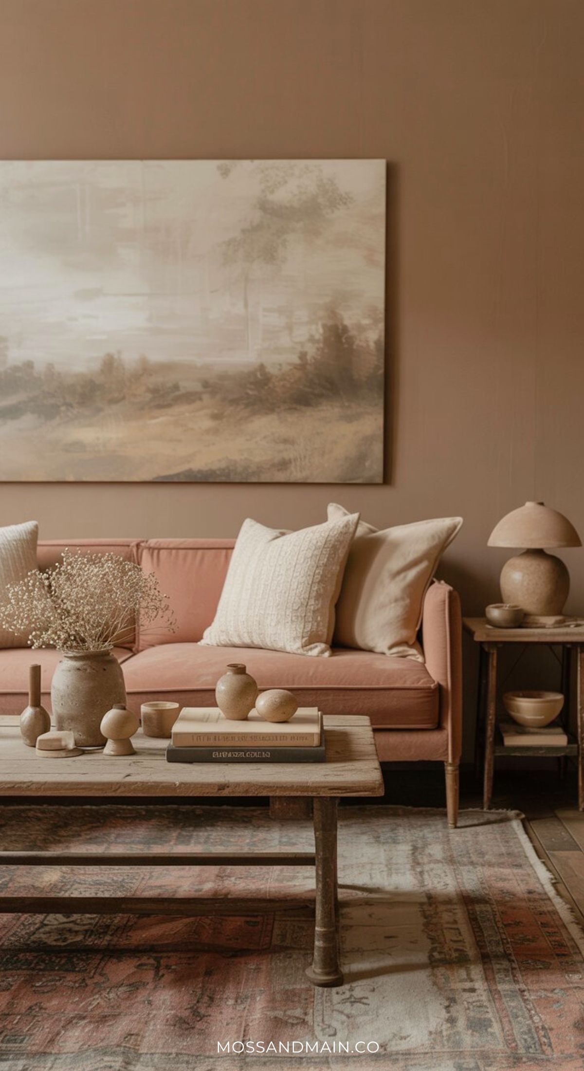 A cozy living room with a rust couch, cream pillows, a rustic coffee table with vases and books, a table lamp, earth-toned decor, and a large landscape painting on a warm beige wall.