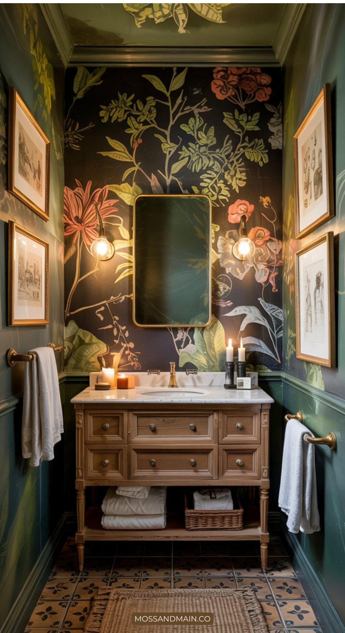 A small, stylish bathroom highlights 2026 Bathroom Trends with dark floral wallpaper, a wooden vanity and marble sink, lit wall sconces beside a rectangular mirror, framed art on green walls, and white towels on gold towel bars.
