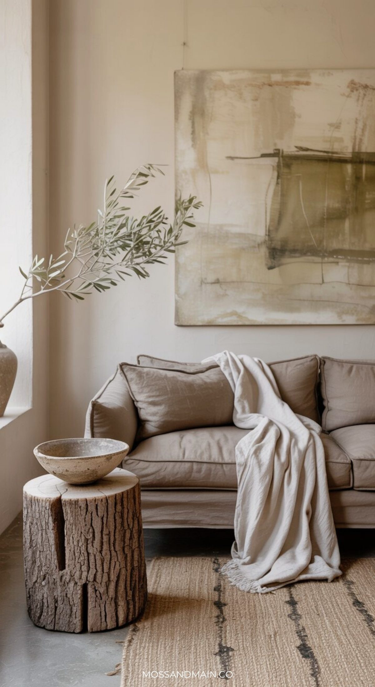 A cozy living room with a beige sofa draped with a light throw, rustic wooden stump side table, textured rug, leafy branches in a vase, and abstract art on the wall—perfect inspiration for earthy living room ideas.