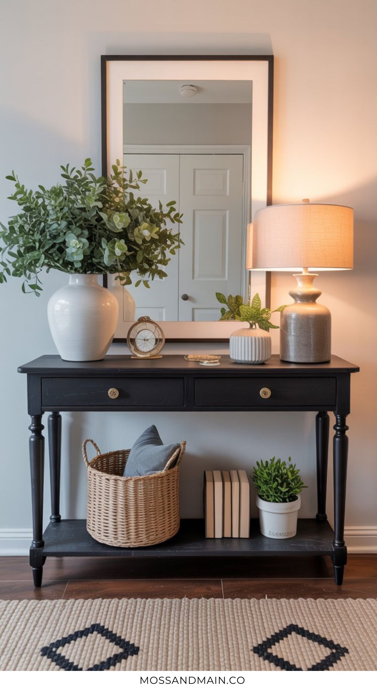 A black entryway table with two drawers holds a white vase with greenery, a lamp, books, clock, and candle. The lower shelf features a basket with a pillow, stacked books, and a potted plant. A mirror completes this entryway table decor.