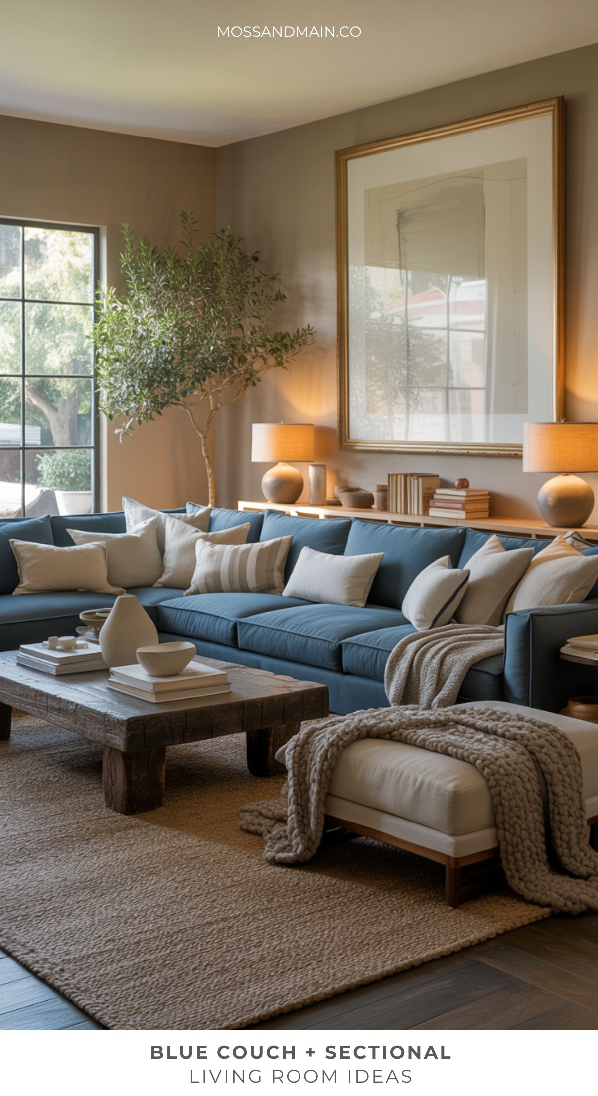 A cozy living room features a Blue couch with neutral pillows, a wooden coffee table, knit throw blanket, table lamps, dramatic framed artwork, and an indoor tree—showcasing inviting living room ideas in warm, natural tones.