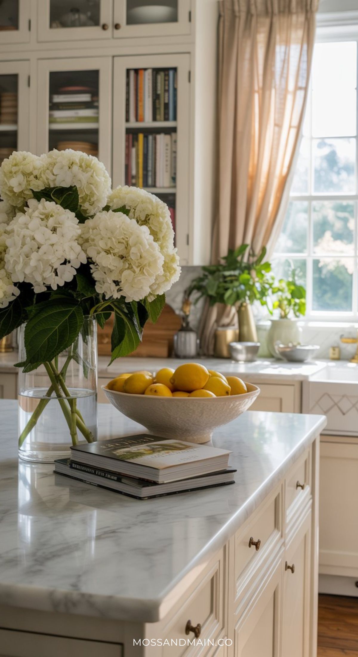 A bright kitchen with white cabinets and marble countertops embodies the Nancy Meyers home aesthetic, featuring a vase of hydrangeas, a bowl of lemons, and stacked books on the island as sunlight streams through sheer curtains.