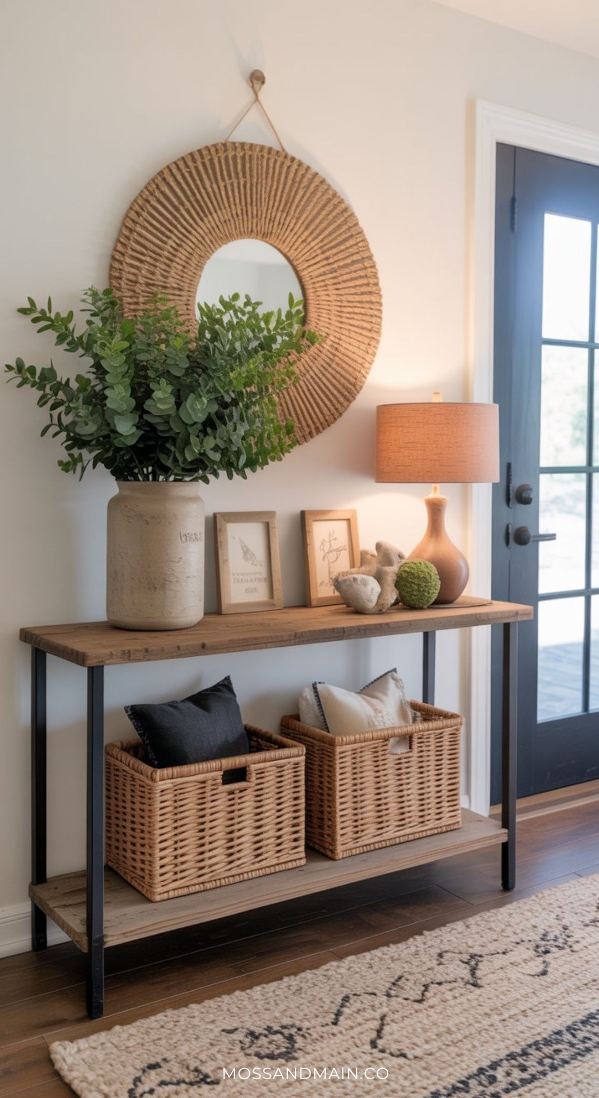 A modern entryway features stylish entryway table decor with a wooden console, potted plant, framed art, a lamp, and baskets. A round woven mirror hangs above the table near a black door, complemented by a patterned rug on the floor.