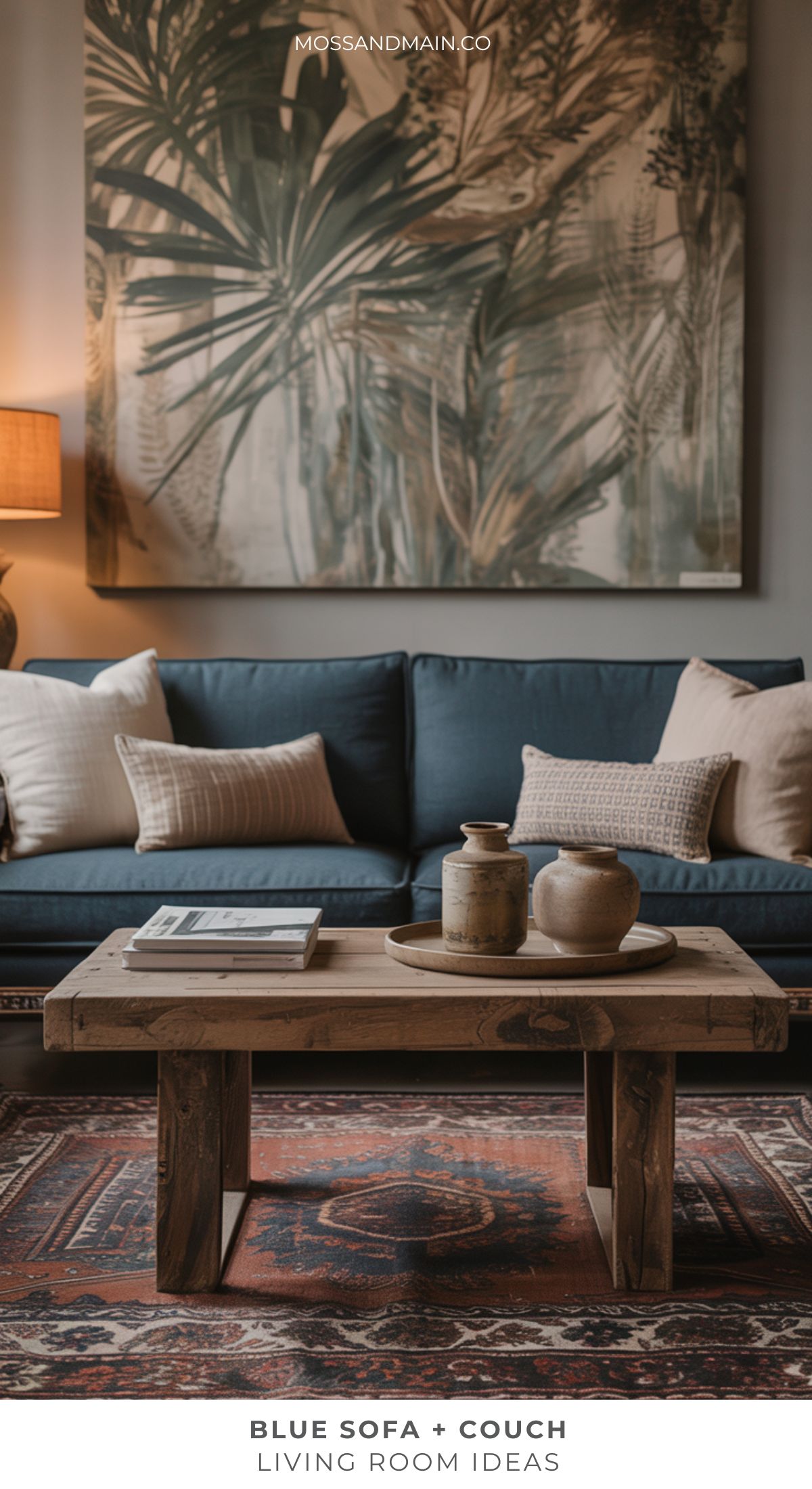 A cozy living room with a Blue couch, beige pillows, a rustic wooden coffee table holding ceramic vases, a patterned rug, table lamp, and large botanical wall art. Text reads: Blue Sofa + Living Room Ideas for stylish home decor.