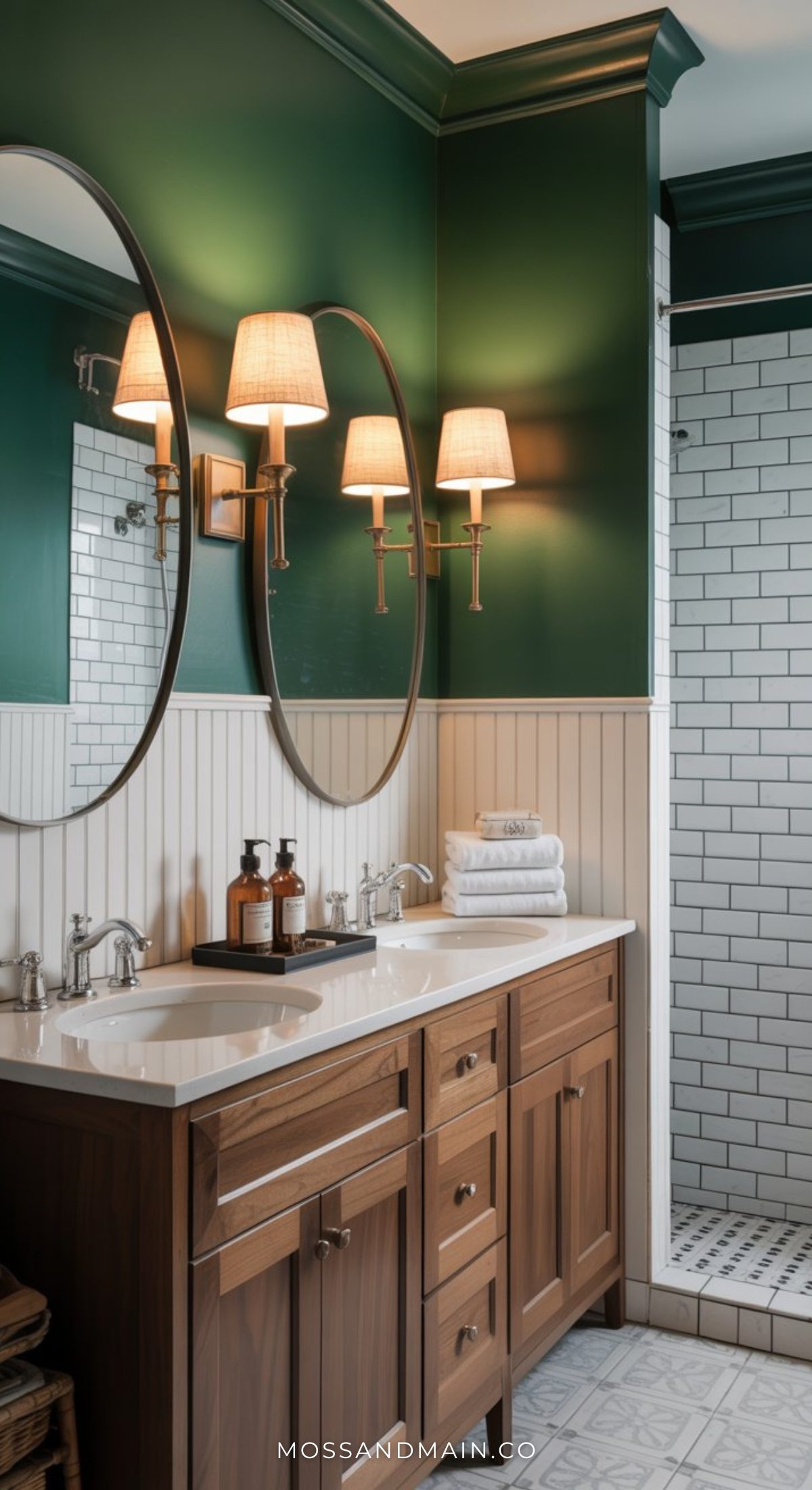 A moody bathroom featuring a wooden double-sink vanity, round mirrors, wall-mounted lamps, dark green upper walls, white paneling, and a tiled walk-in shower. Towels and toiletries sit on the counter.