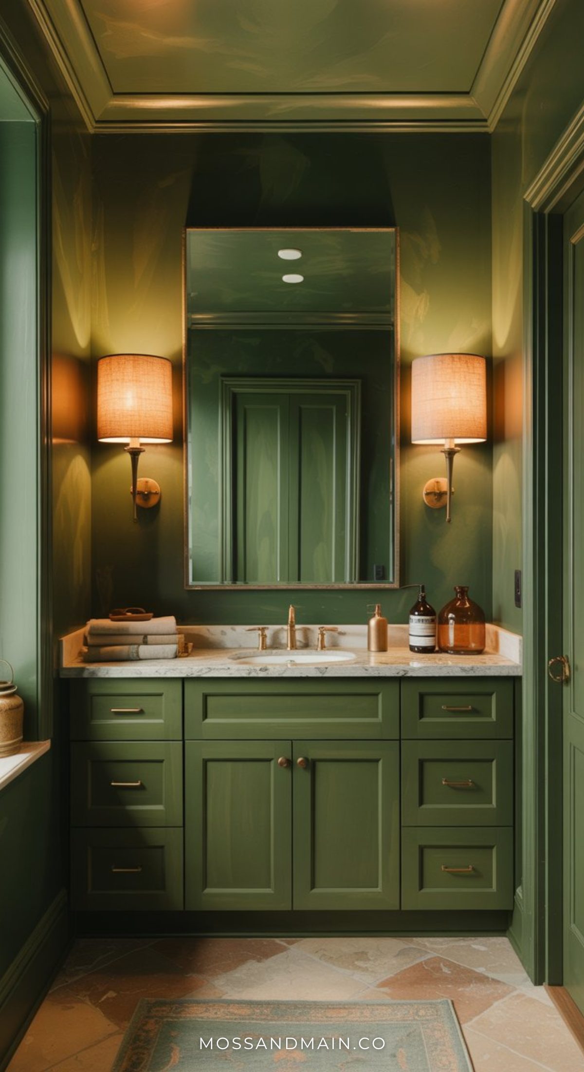 A luxe bathroom featuring dark green cabinetry, a white marble countertop, brass fixtures, and two wall sconces with beige shades flanking a large mirror. Neutral decor accents sit elegantly on the counter.