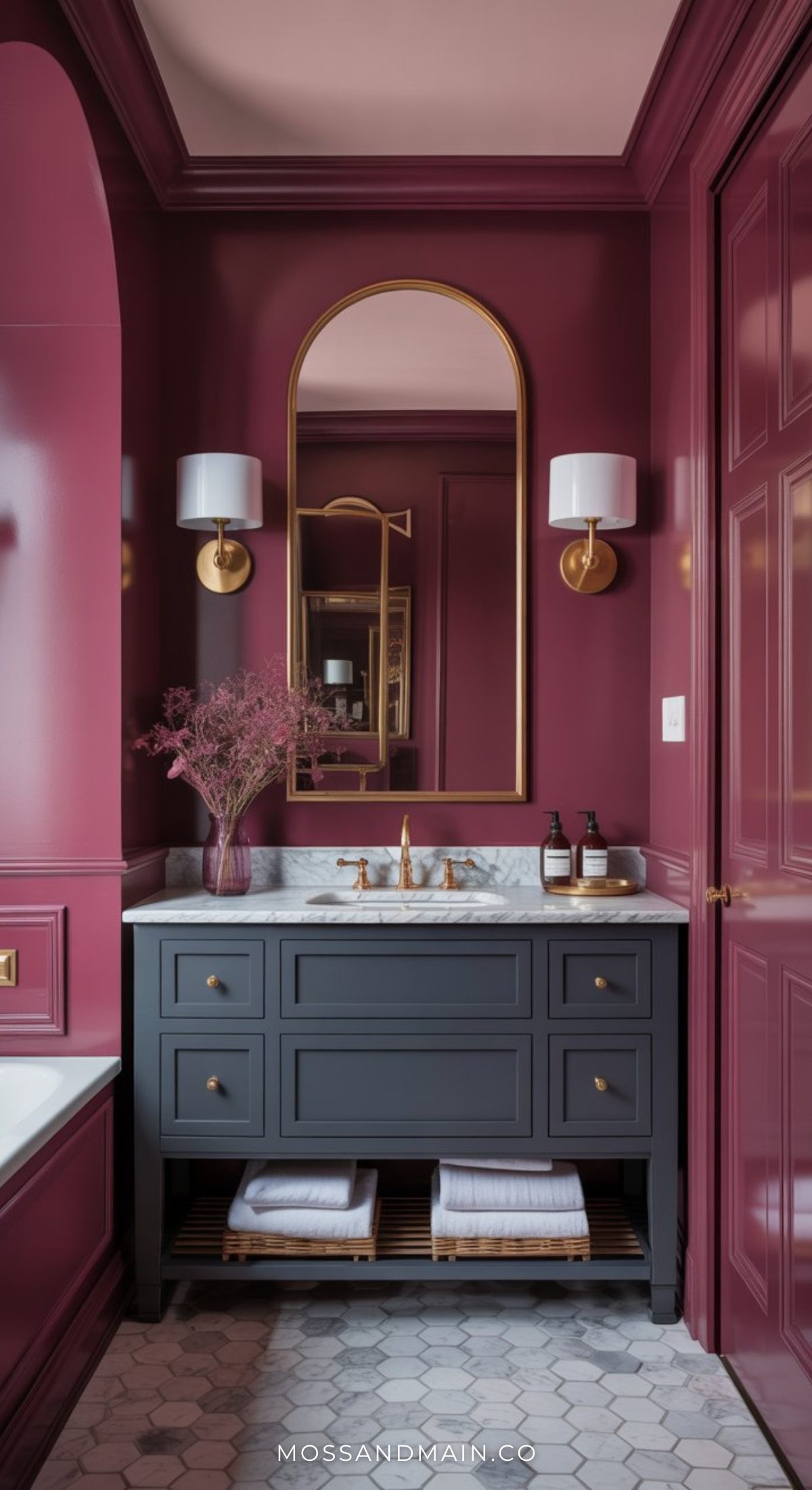 A stylish bathroom with plum-colored walls, a gold-framed mirror, marble countertop, and navy blue vanity with brass hardware showcases bold bathroom decor, complemented by white sconces and towels neatly stacked on a wooden shelf below.